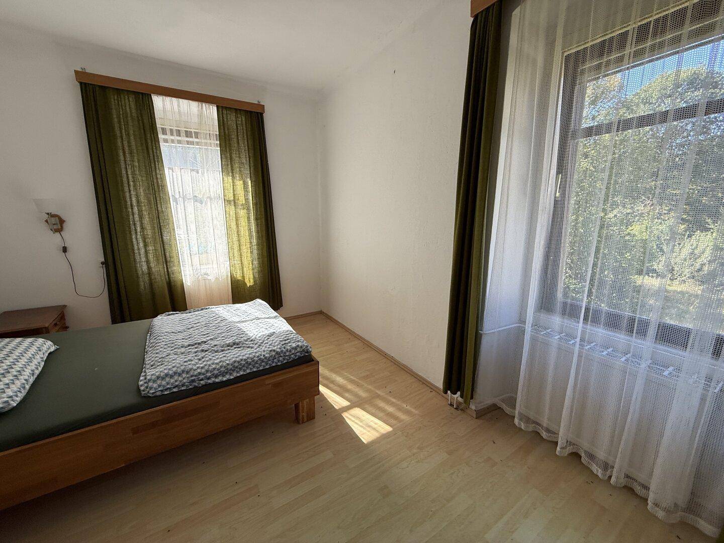 Schlafzimmer mit Holzbett und zwei Fenstern, die viel Tageslicht hereinlassen.