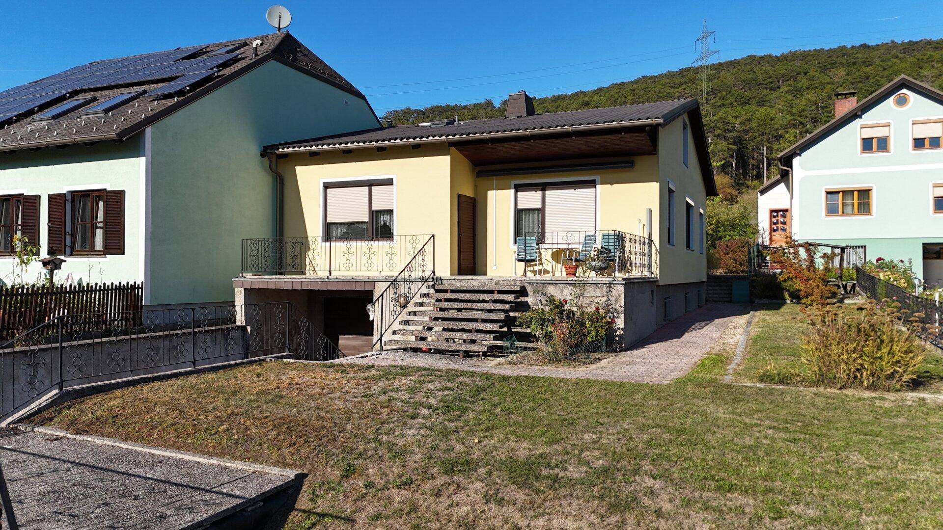 Gelbes Wohnhaus mit angrenzendem Garten und Blick auf die bewaldeten Hügel.