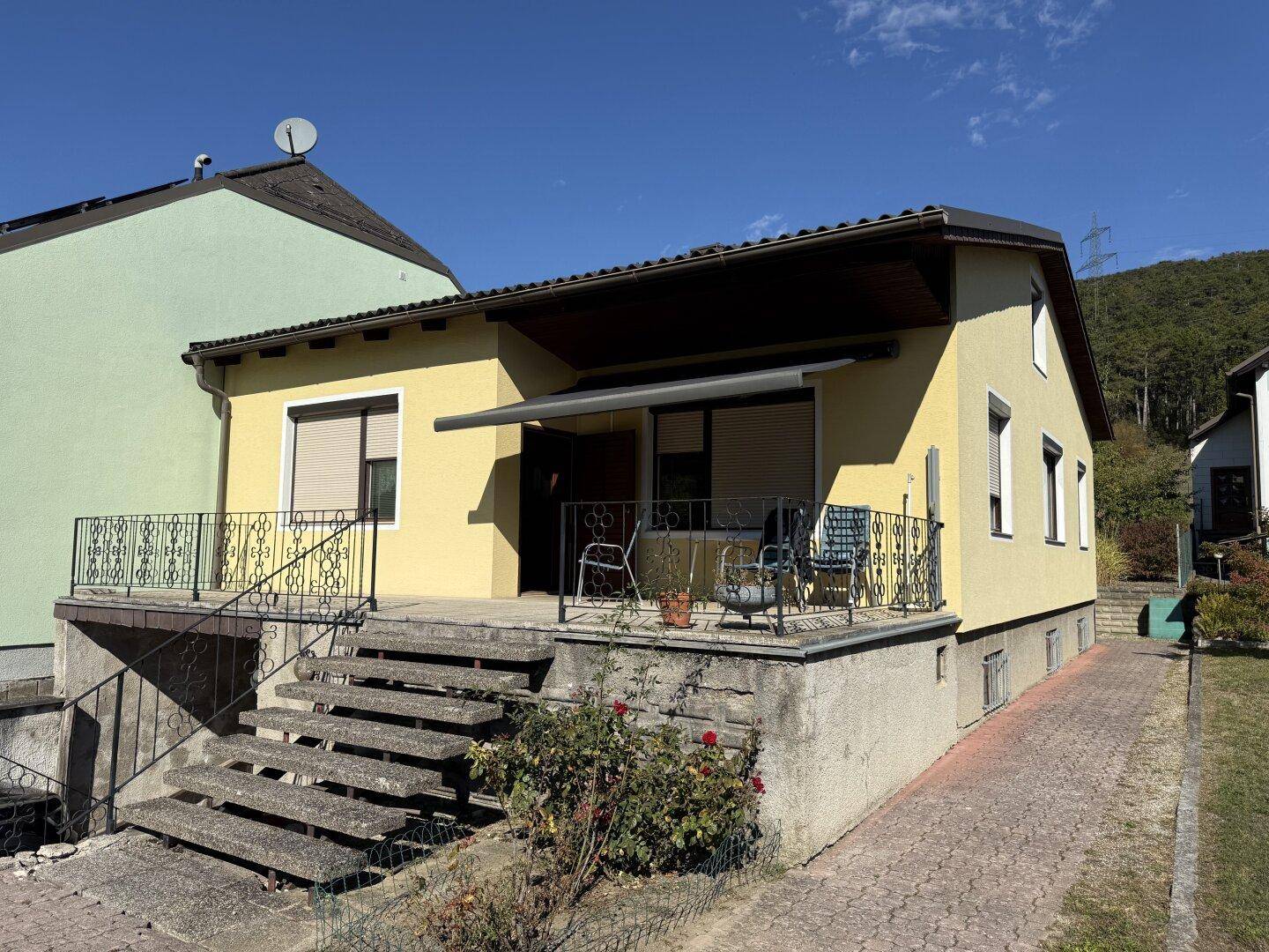 Vorderansicht des gelben Hauses mit kleiner Terrasse und Treppe unter blauem Himmel.