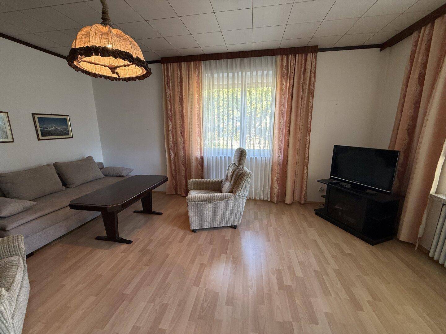 Helles Wohnzimmer mit Laminatboden, Sofa, Sessel und Fernseher.