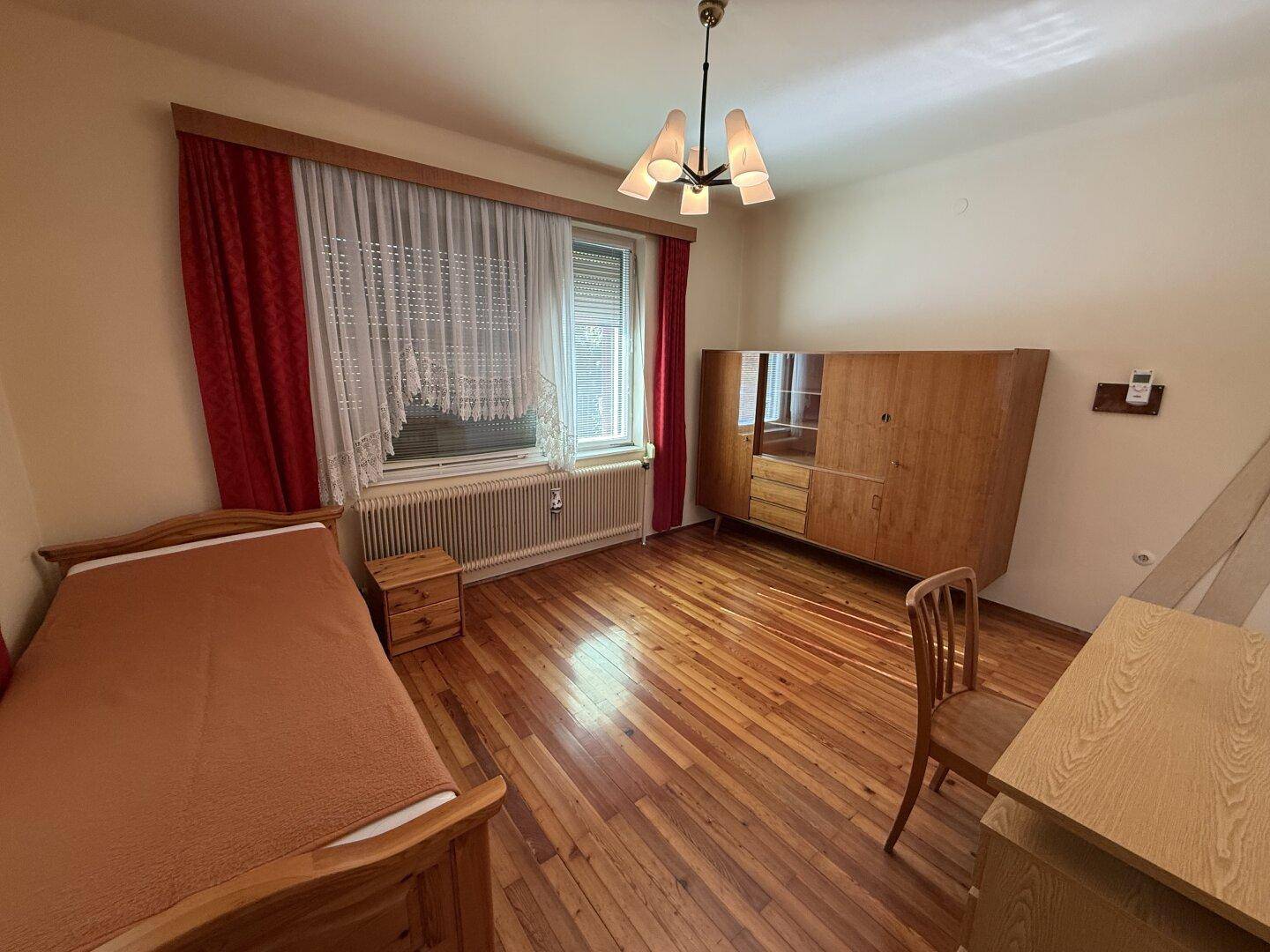 Schlafzimmer mit Holzboden, Einzelbett, Schrank und Fenster mit rotem Vorhang.