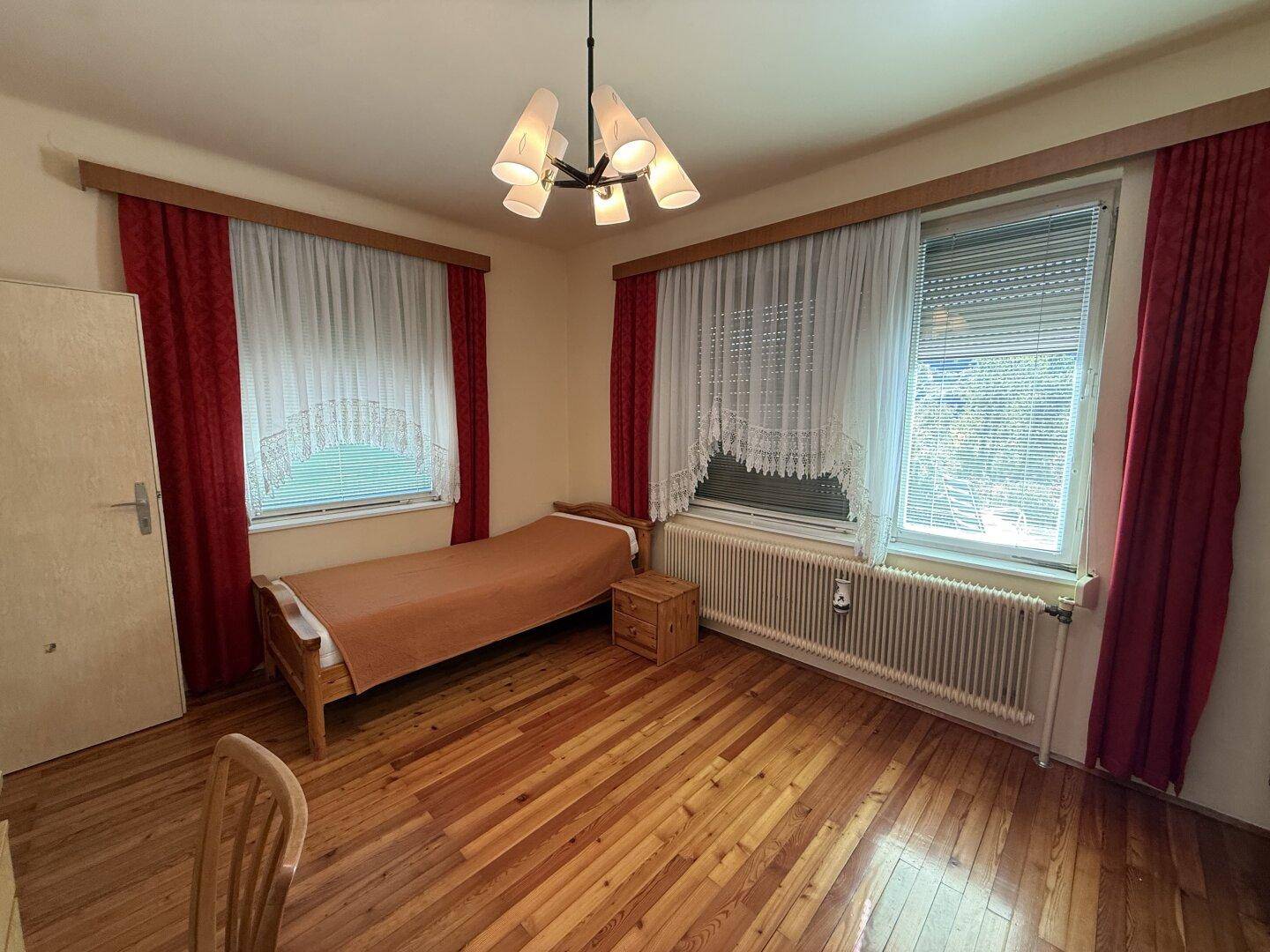 Helles Schlafzimmer mit Holzboden, Einzelbett und zwei Fenstern mit Vorhängen.