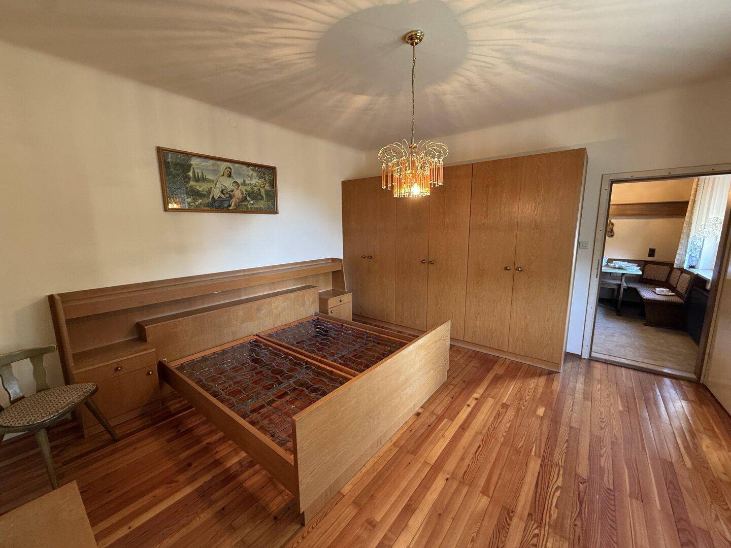 Schlafzimmer mit Holzboden, Doppelbett, Kleiderschrank und Zugang zu einem weiteren Raum.