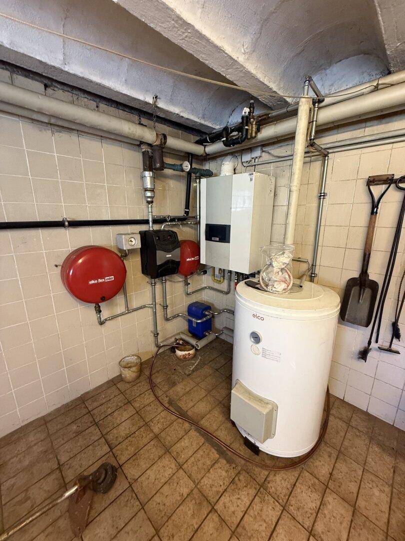 Technikraum im Keller mit Heizungsanlage, Warmwasserspeicher und sichtbaren Rohrleitungen.