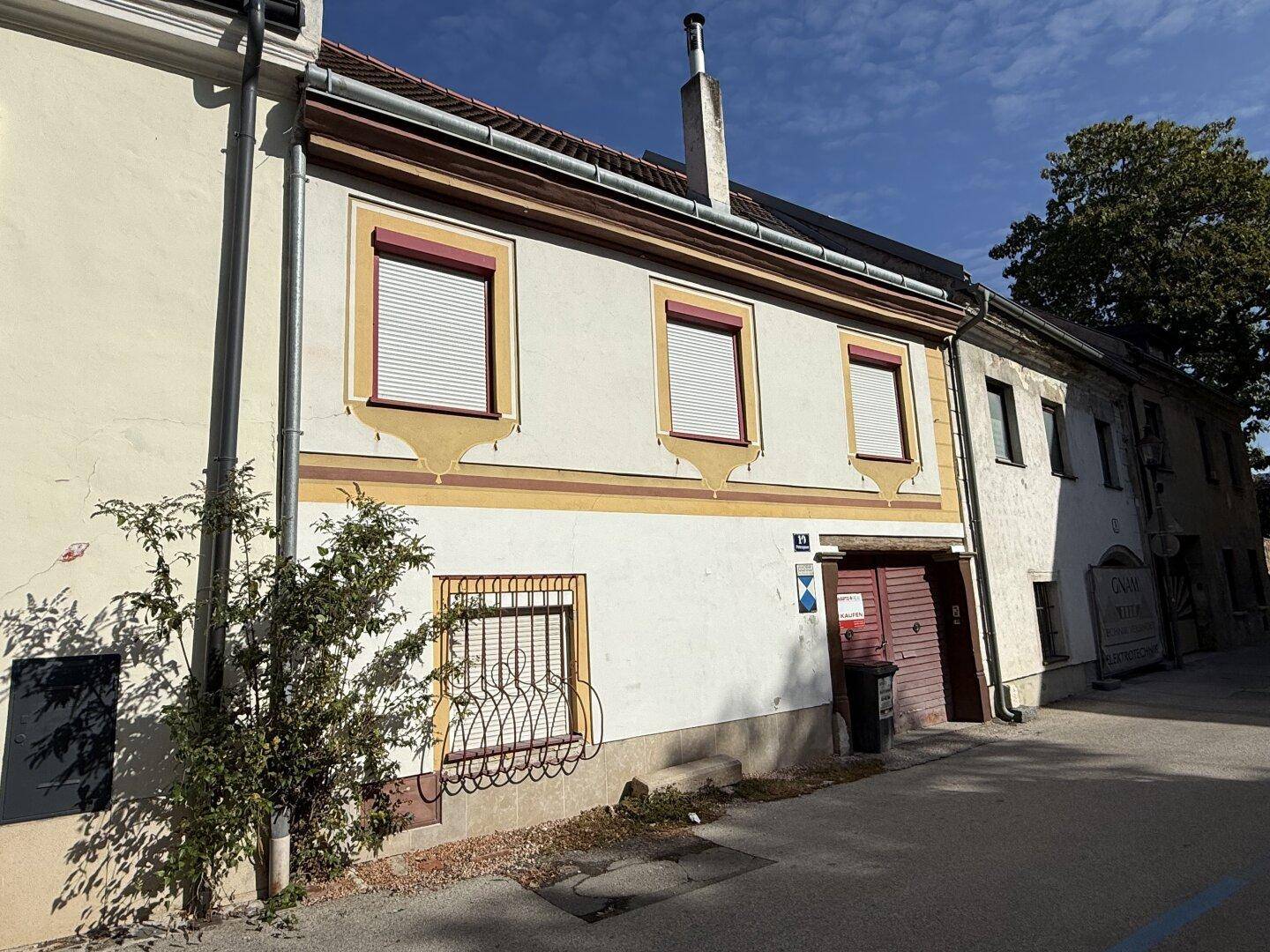 Seitenansicht des Gebäudes mit traditioneller Architektur, roten Fensterläden und einem Baum im Hintergrund.