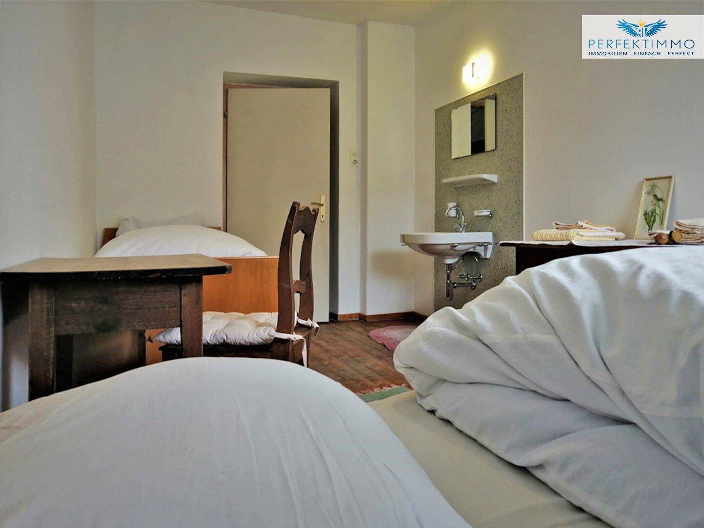 Schlafzimmer mit Einzelbetten, Holzboden und einem kleinen Waschbeckenbereich an der Wand.