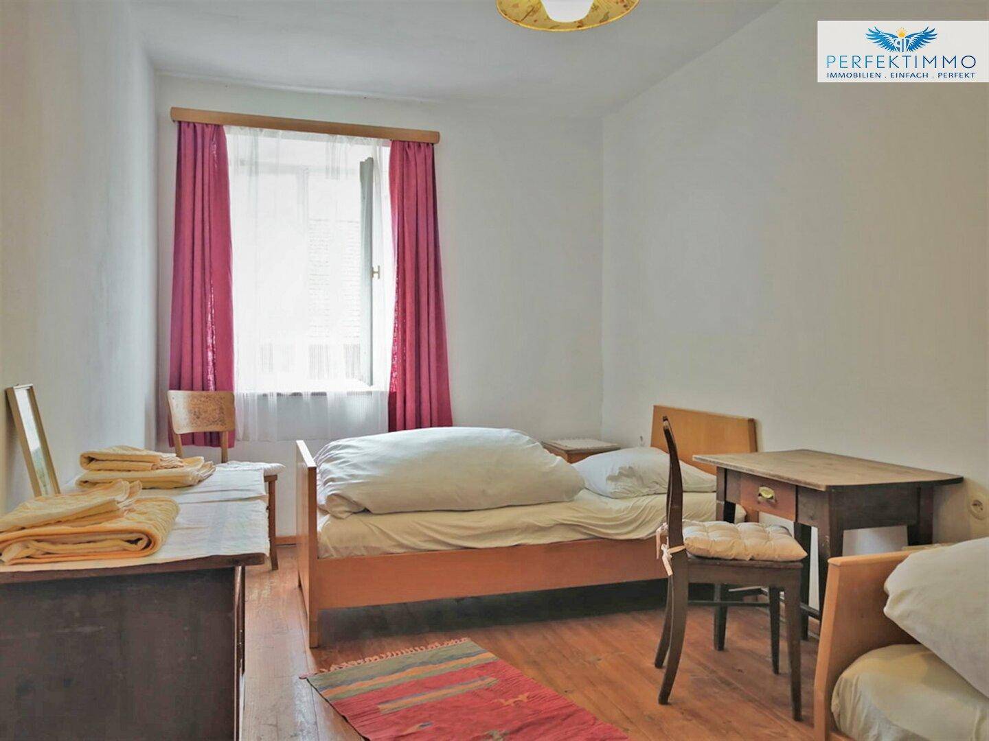 Einfaches Schlafzimmer mit zwei Betten, Holzmöbeln und einem Fenster mit rotem Vorhang.