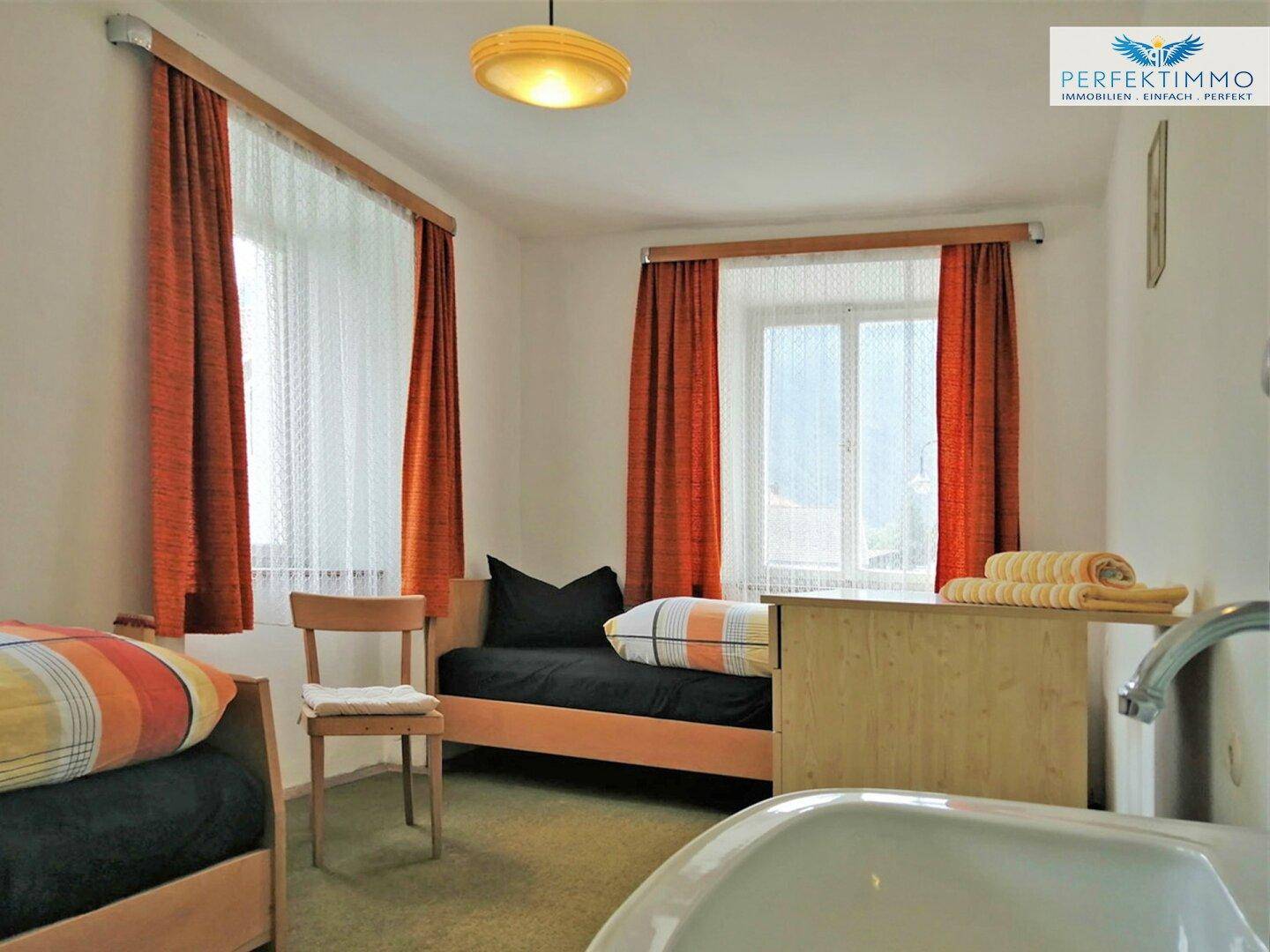 Helles Schlafzimmer mit zwei Betten, Teppichboden und einem Fenster mit orangefarbenen Vorhängen.