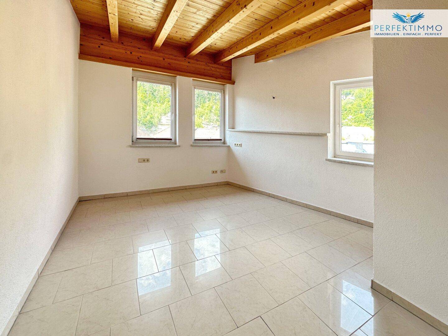 Heller Raum mit sichtbaren Holzbalken an der Decke, Fliesenboden und grüner Aussicht.