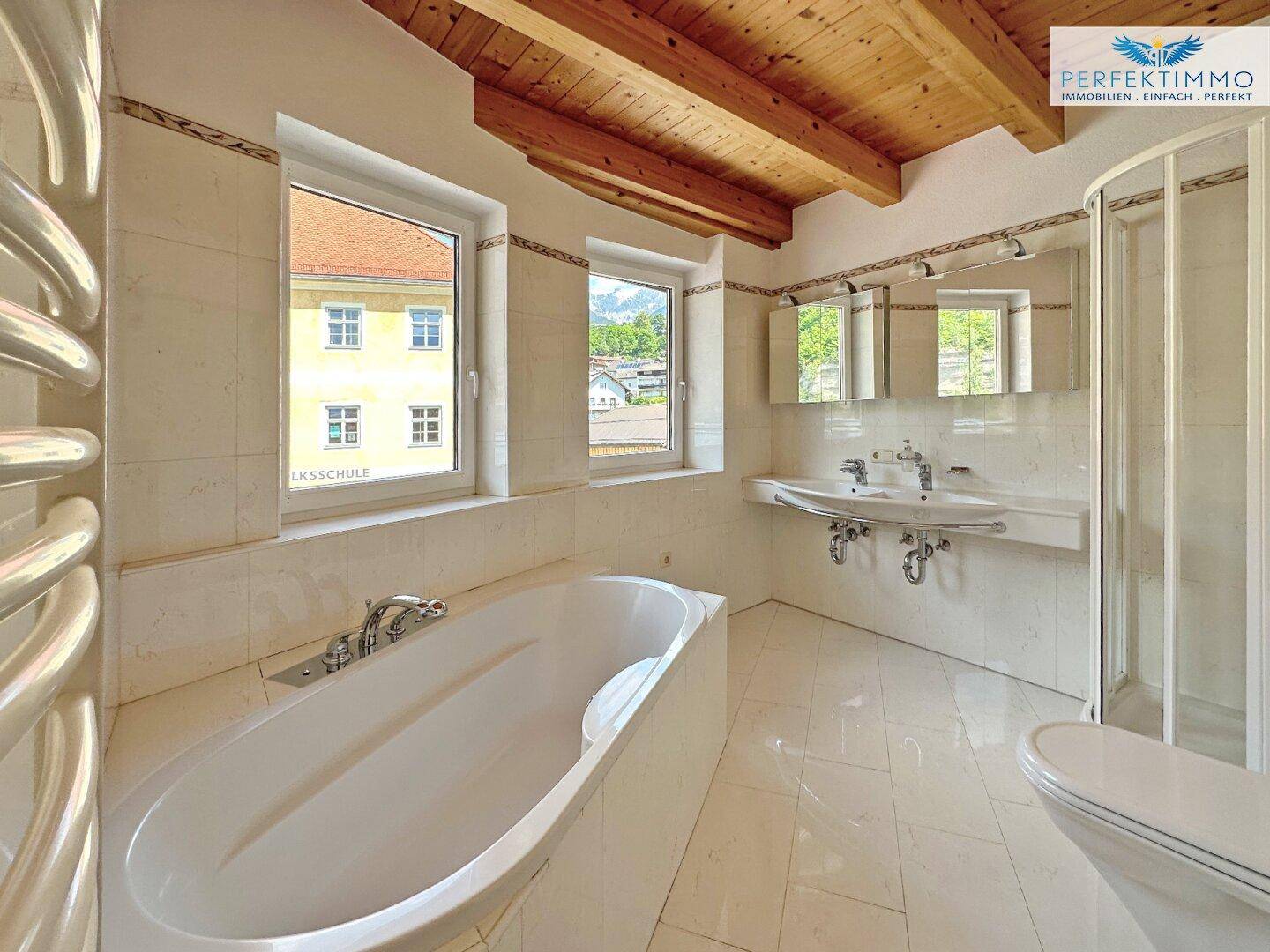 Helles Badezimmer mit Badewanne, Doppelwaschbecken, Duschkabine und Holzbalkendecke.