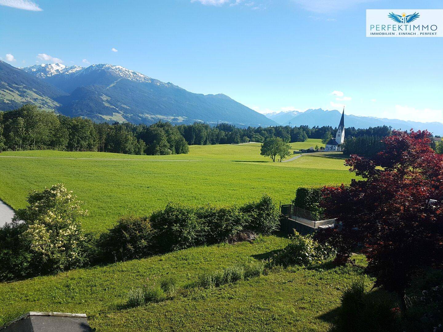 Weitläufiger Ausblick über grüne Wiesen, Wälder und eine Kirche mit majestätischen Bergen im Hintergrund.