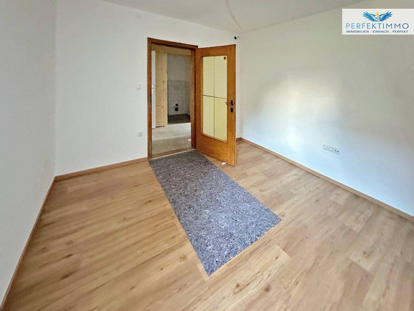 Leerer Raum mit Laminatboden und Holztür, der zu einem weiteren Bereich führt.