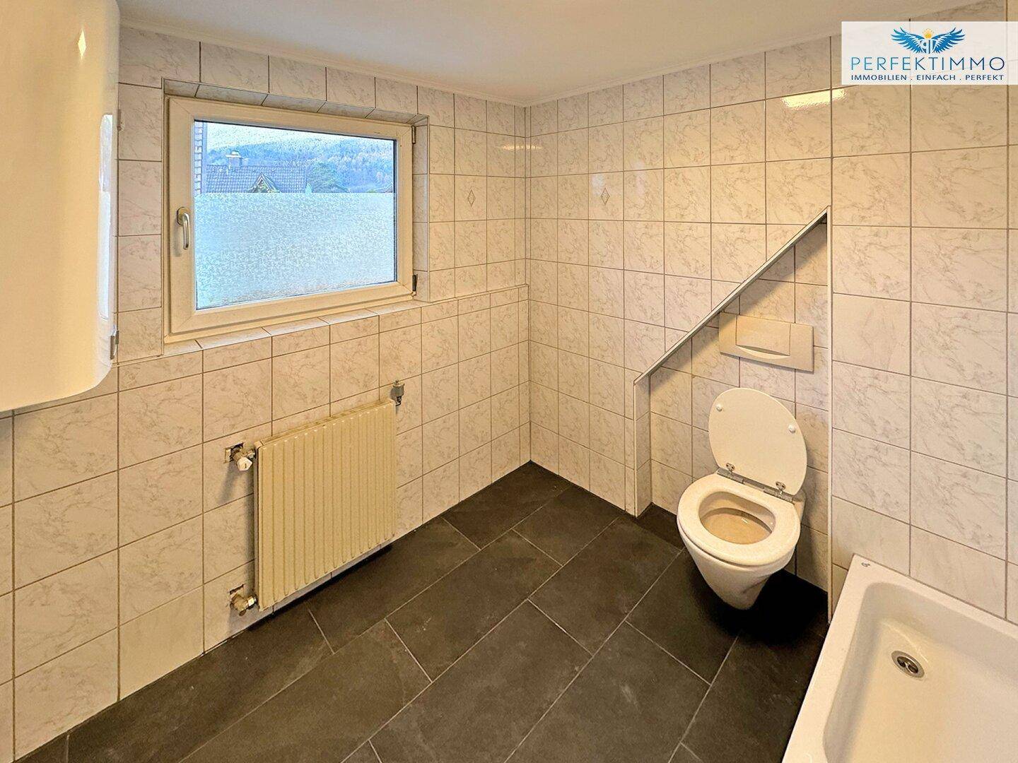 Geflieste Toilette mit WC, Waschbecken, Fenster und Warmwasserboiler.