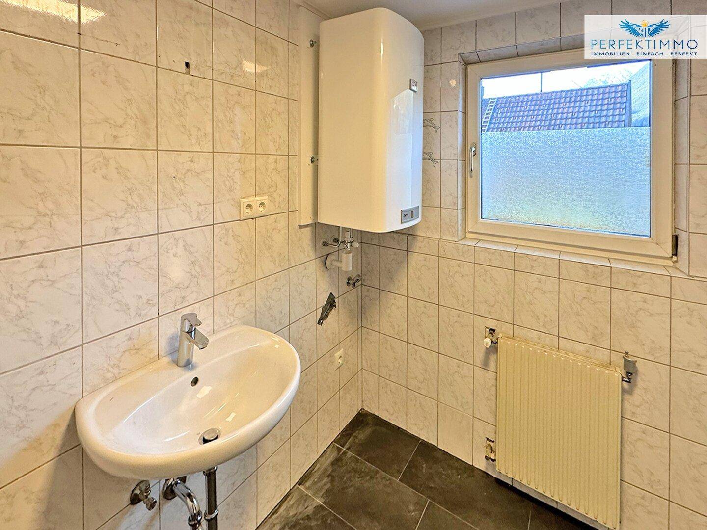 Gefliestes Badezimmer mit Waschbecken, Fenster und Warmwasserboiler.