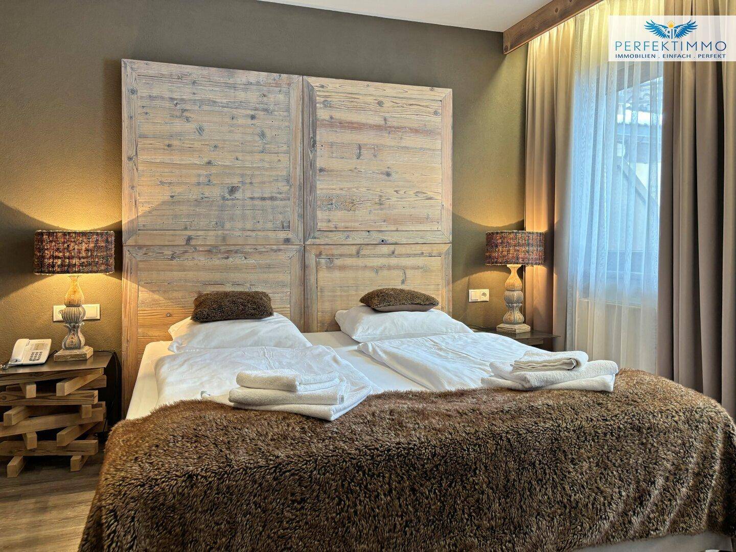 Helles Schlafzimmer mit hellem Holz-Kopfteil, bequemen Betten und Vorhängen am Fenster.