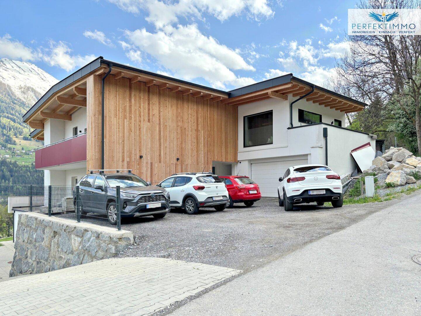 Ansicht des modernen Gebäudes mit Garagenzufahrt und Außenstellplätzen vor alpiner Kulisse.