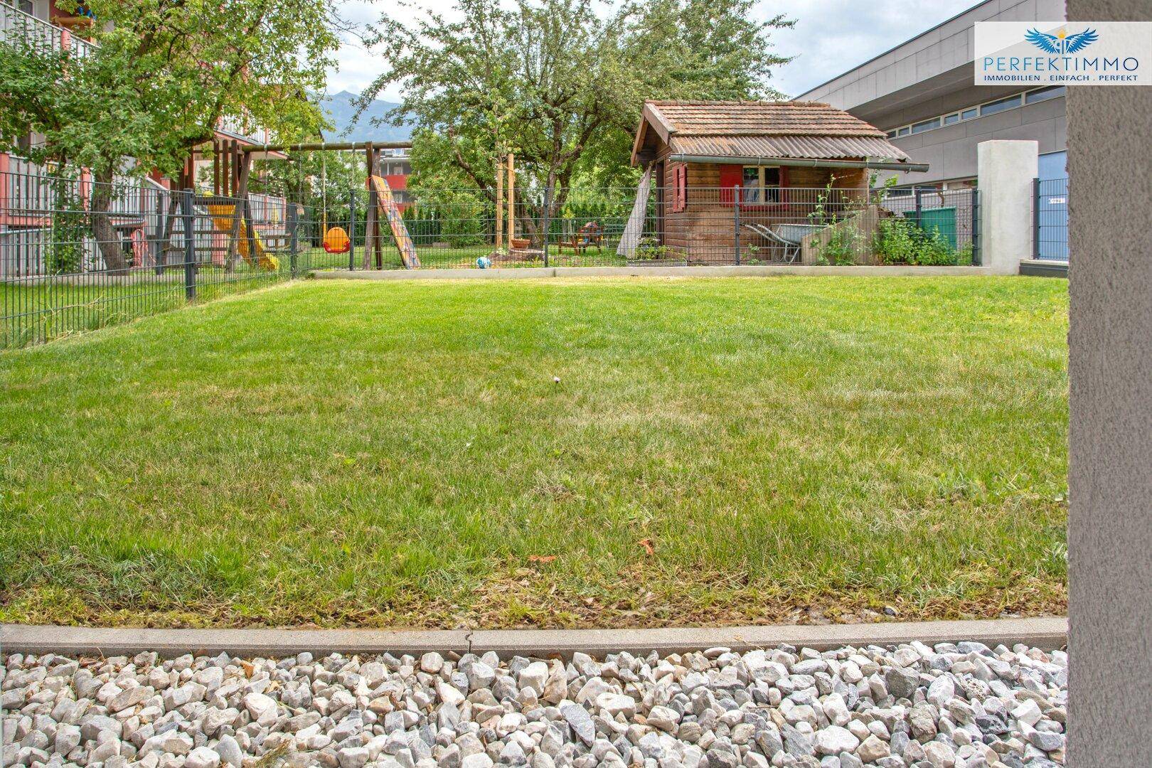 Großer Gemeinschaftsgarten mit Rasenfläche und Kinderspielplatz im Hintergrund.