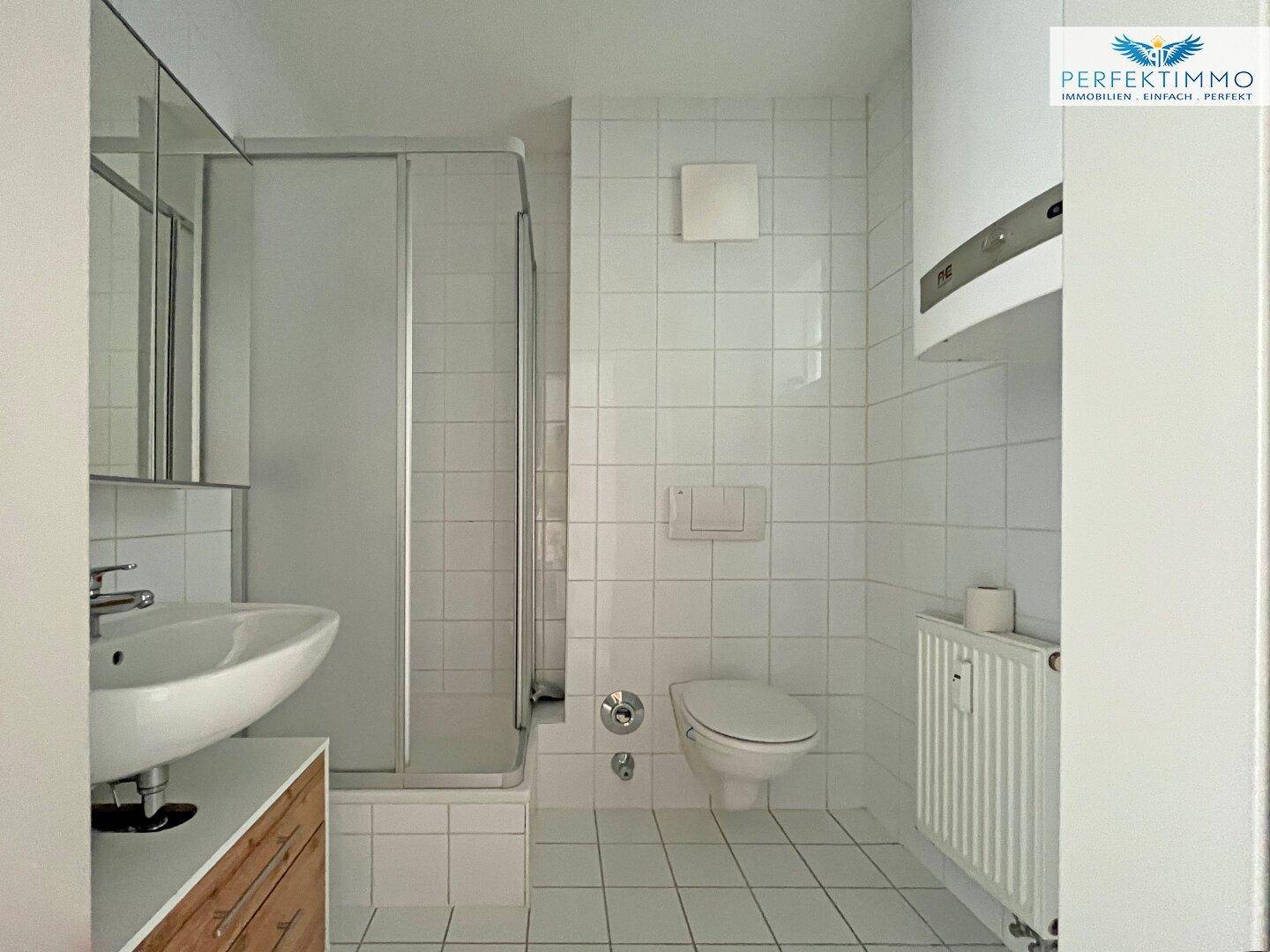 Helles Badezimmer mit weißen Fliesen, Dusche, wandhängendem WC und Heizkörper.