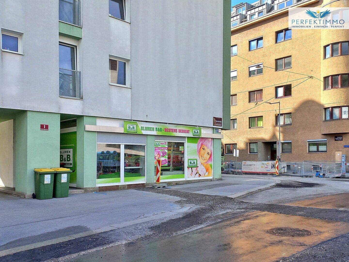 Gebäudeansicht mit Blumengeschäft im Erdgeschoss, umgeben von weiteren Gebäuden und einer Straße.