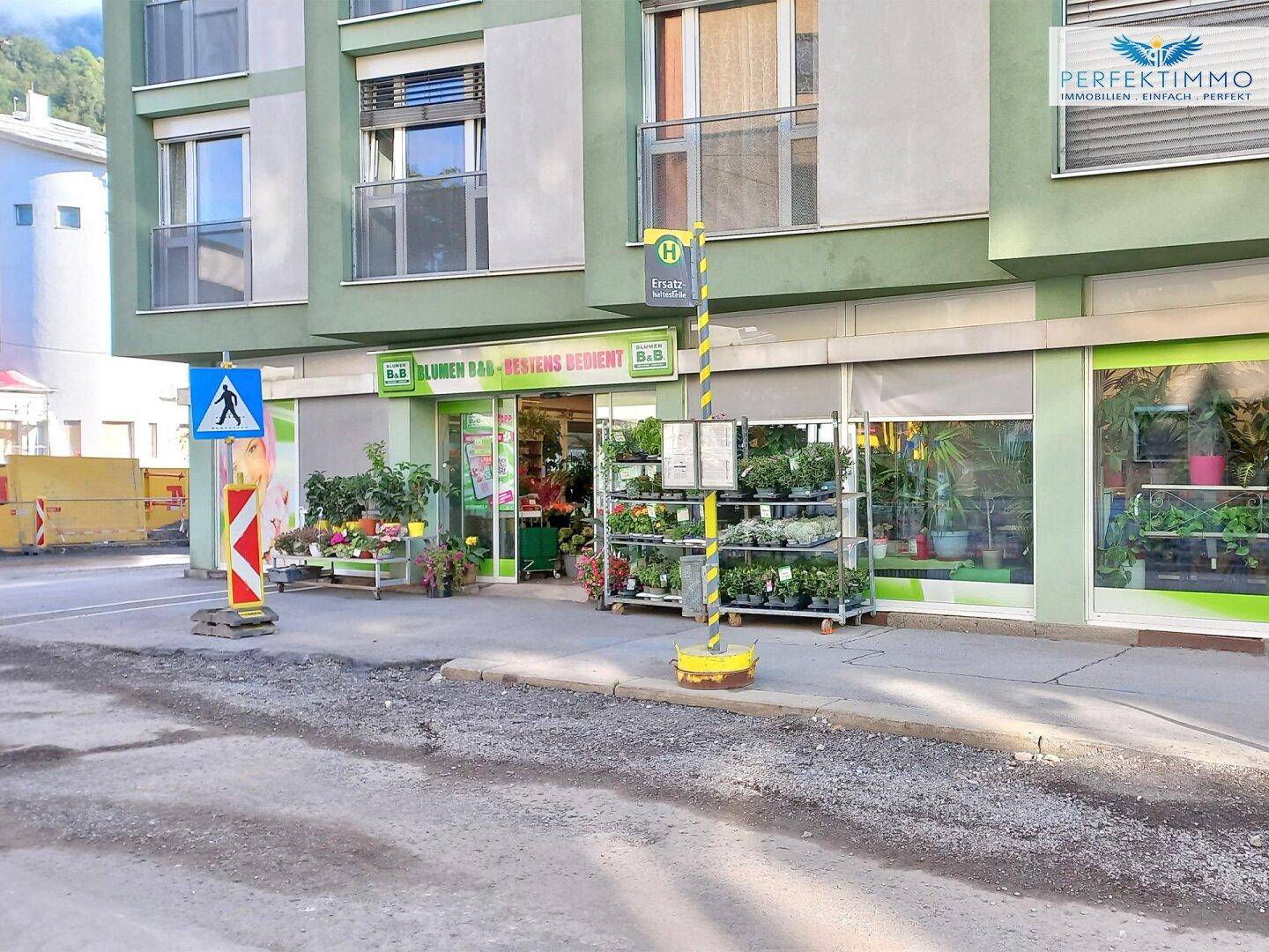 Blumengeschäft an einer Straße mit Bushaltestelle und Pflanzen vor dem Eingangsbereich des Gebäudes.