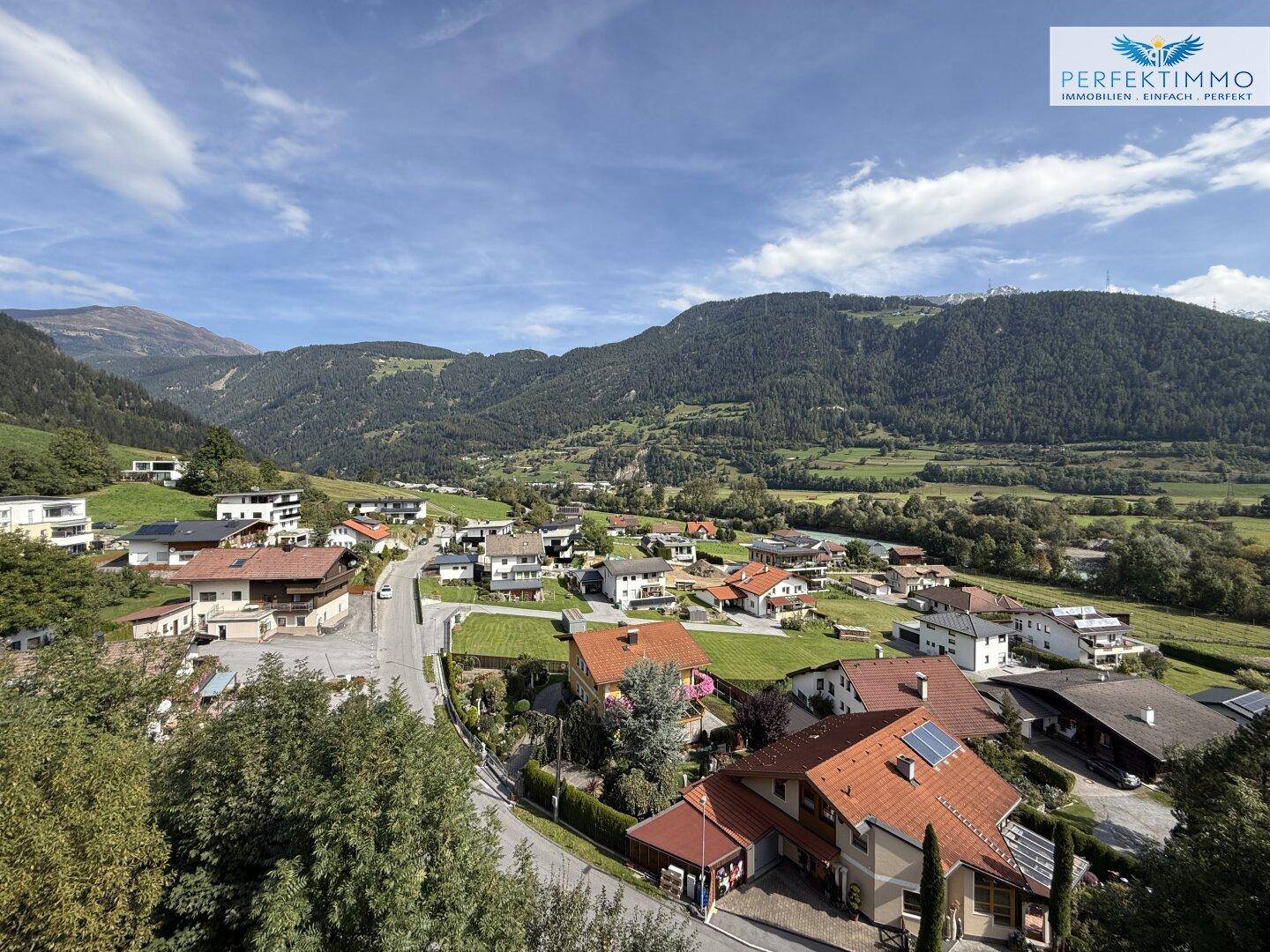 Weitläufiger Blick über eine Siedlung im Tal, umgeben von grünen Hügeln und Bergen.