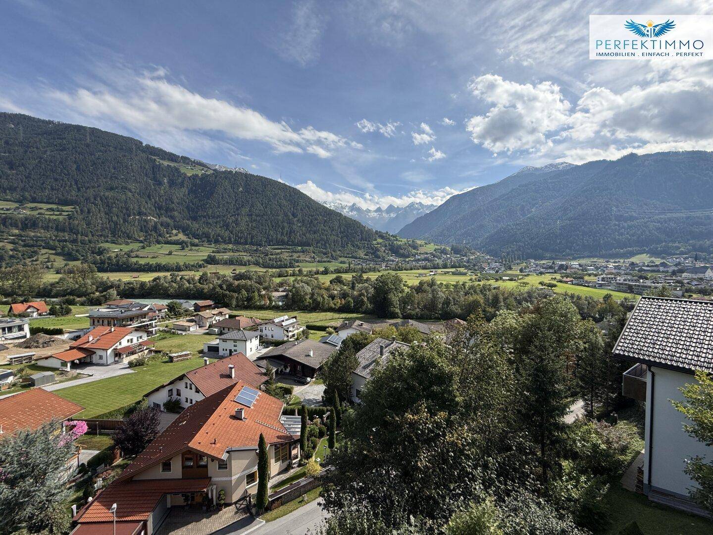 Panoramablick auf ein malerisches Tal mit Häusern und majestätischen Bergen unter blauem Himmel.