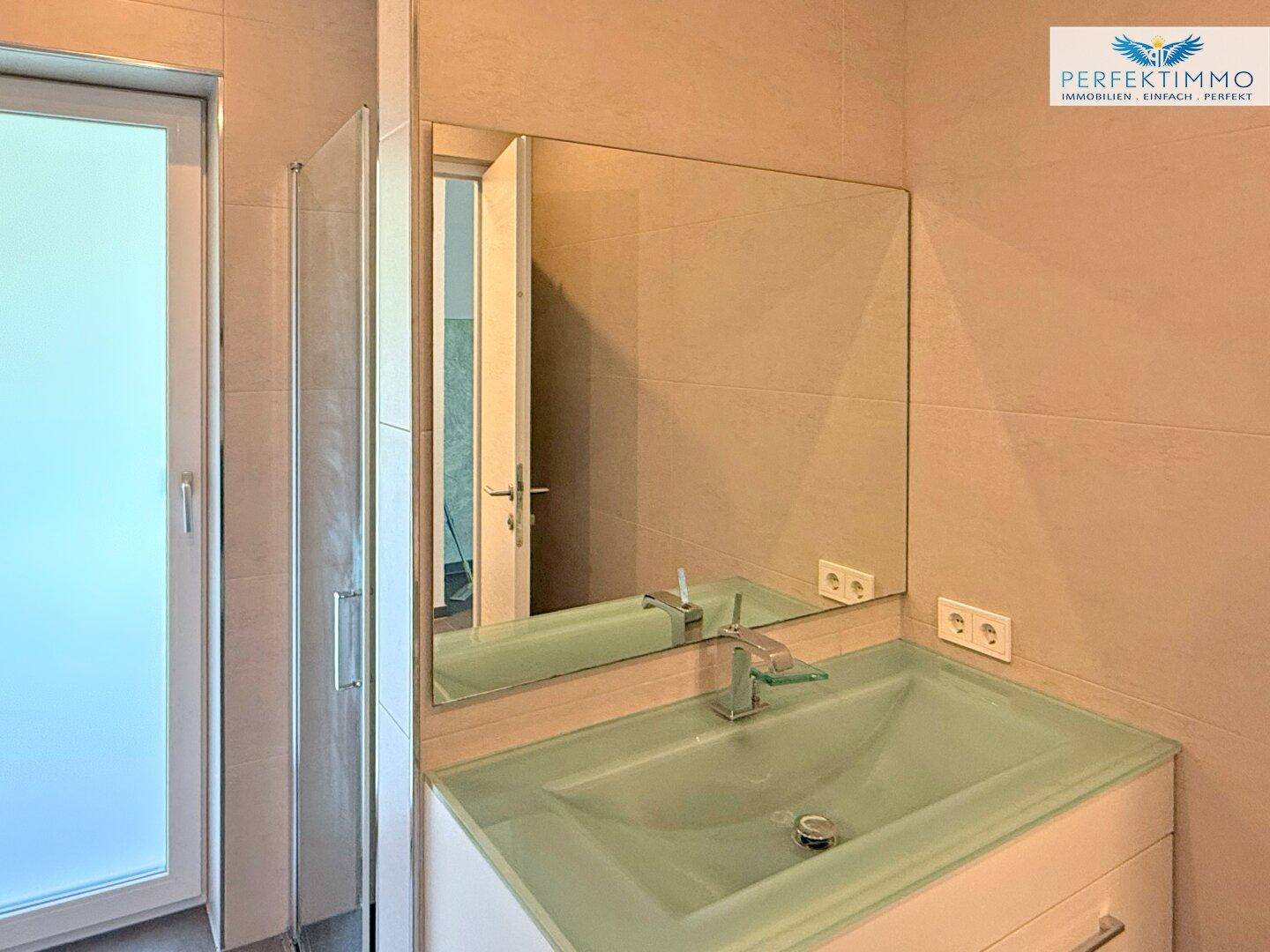 Modernes Badezimmer mit Waschtisch, großem Spiegel und Duschkabine, in hellen Farbtönen gehalten.