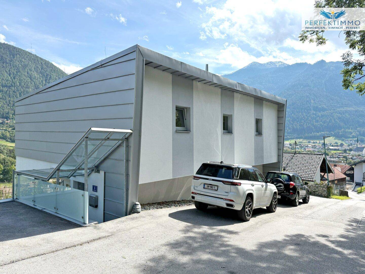 Moderne Gebäudefassade mit einem externen Glaslift und Parkplätzen vor dem Haus.