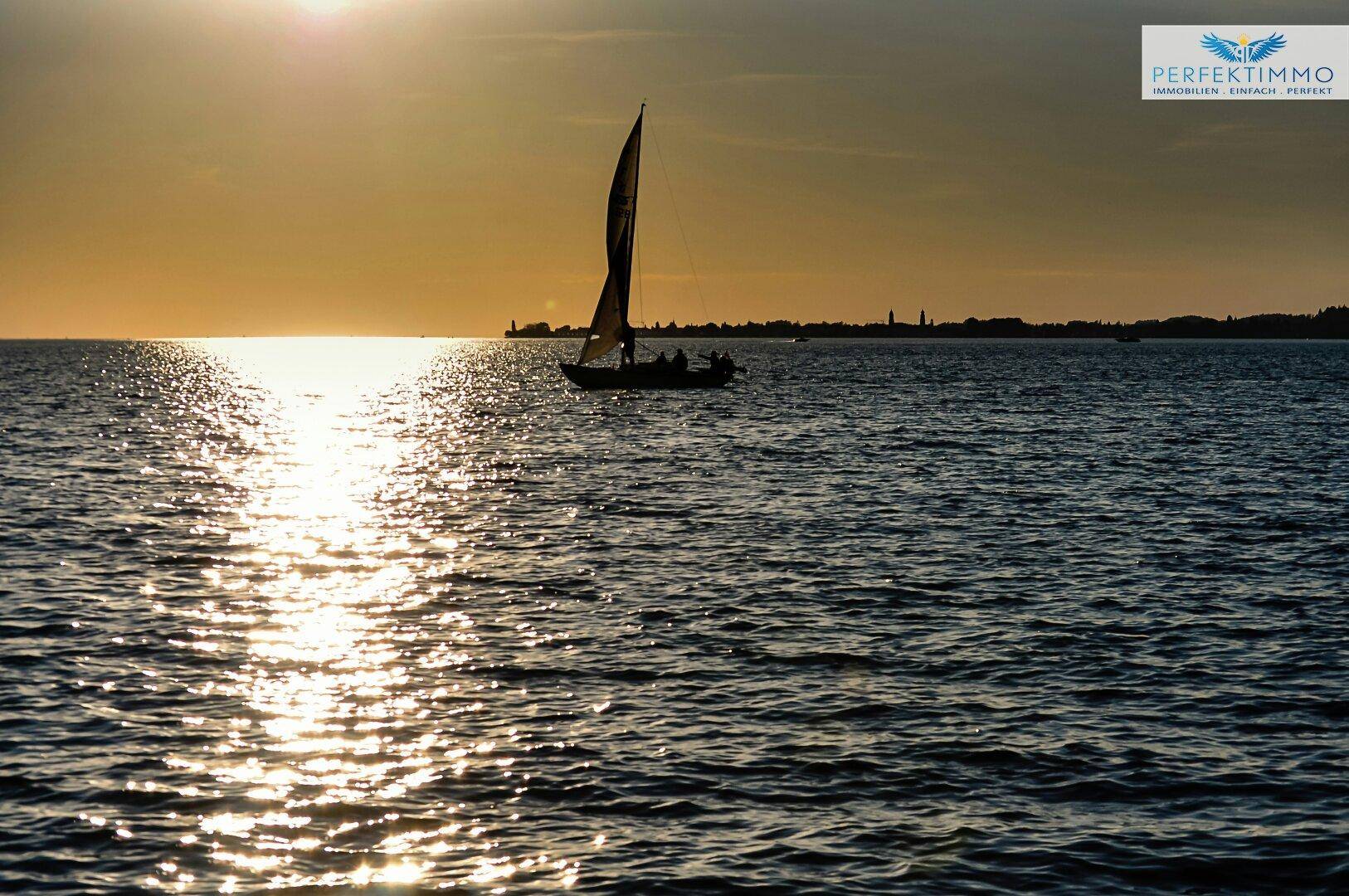 Ein Segelboot auf dem See bei Sonnenuntergang, das Wasser glitzert im goldenen Licht.
