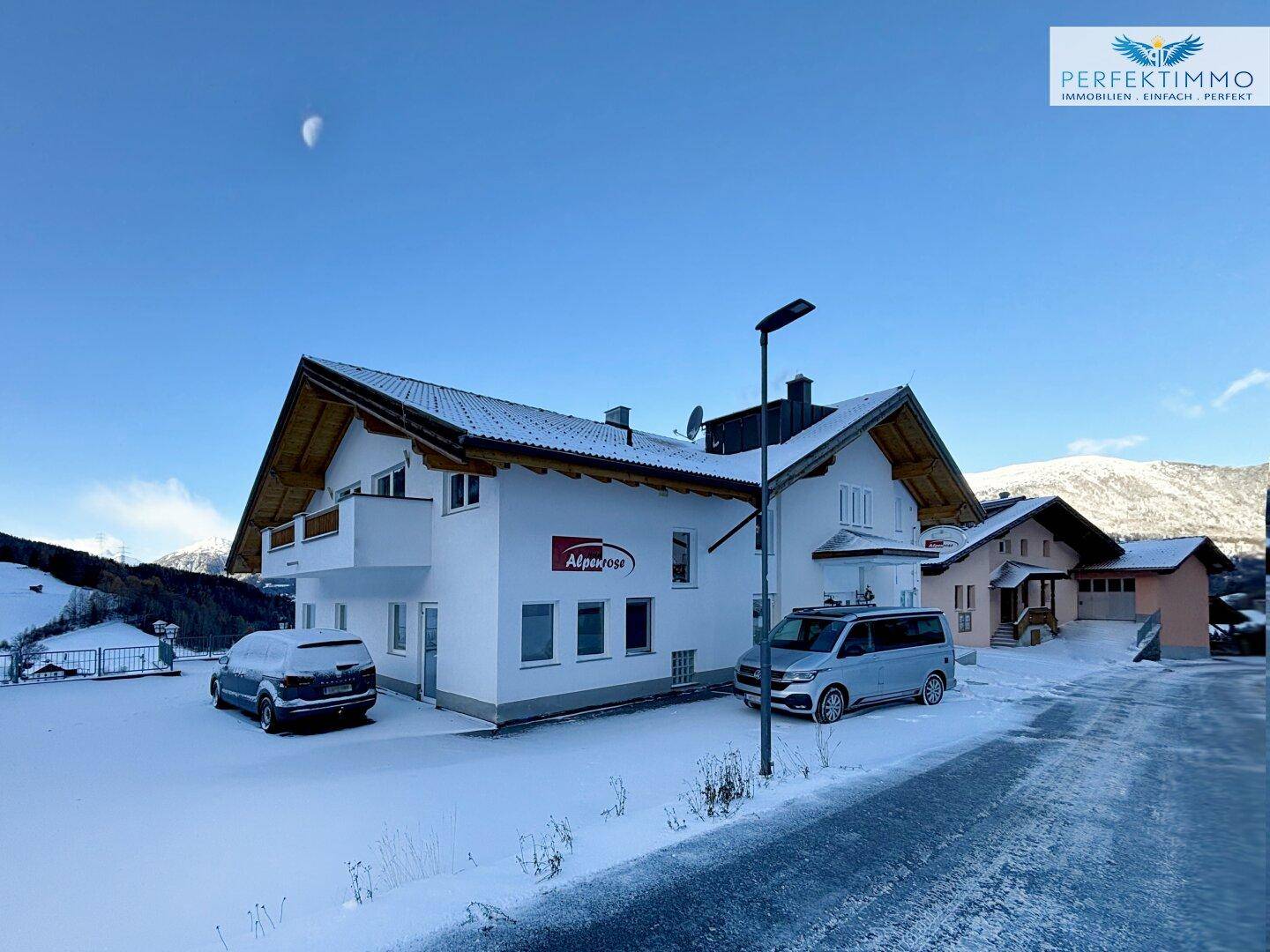 Weißes Gebäude mit Holzbalkonen und Giebeldach, umgeben von Schnee und Bergen unter blauem Himmel.