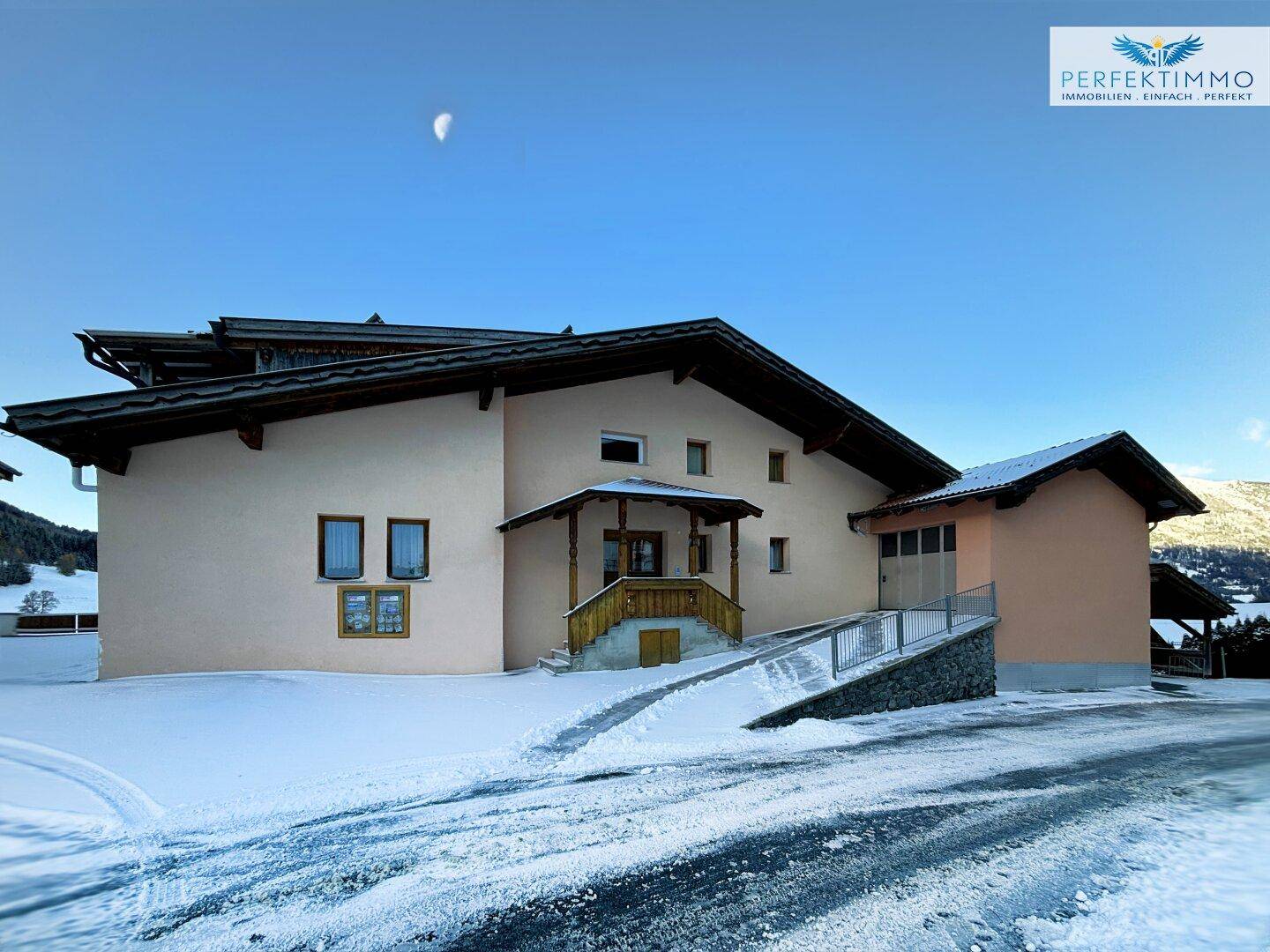 Gebäude mit beiger Fassade und Holzelementen, umgeben von Schnee und einer Zufahrt.