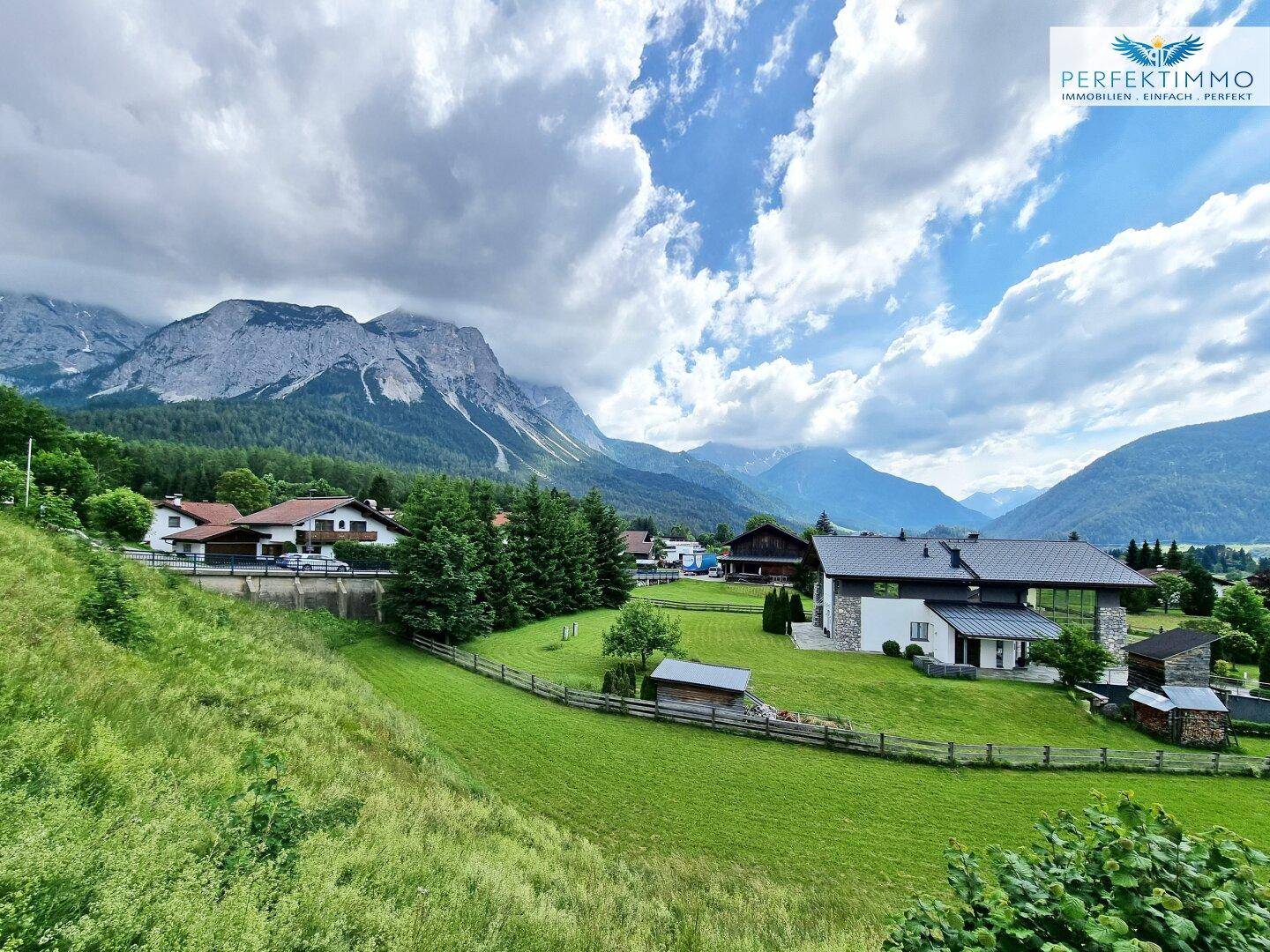 Weitläufige Landschaft mit Häusern, grünen Wiesen und majestätischen Bergen unter blauem Himmel.