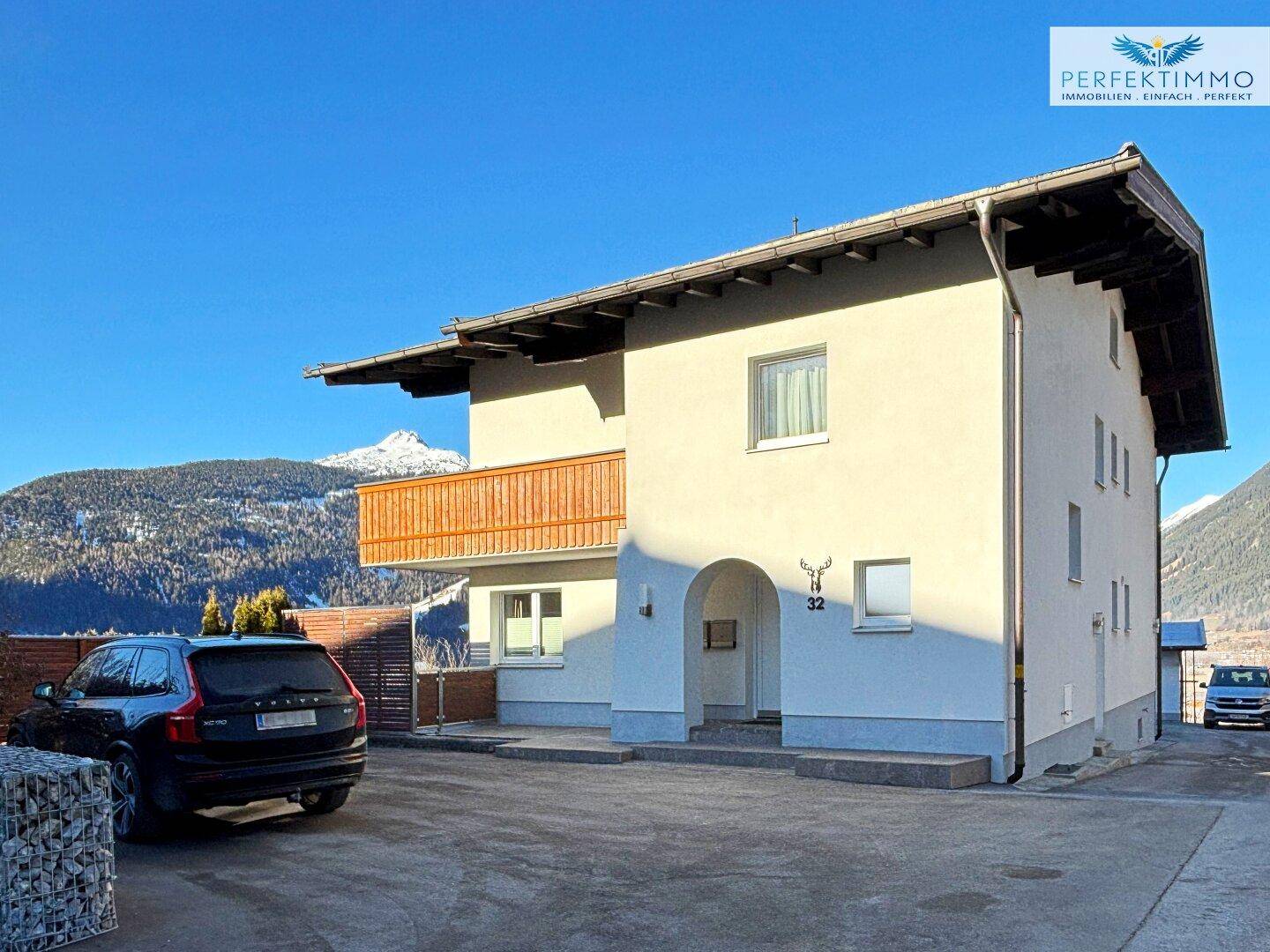 Weiß verputztes Haus mit Holzbalkon und Parkplatz im Vordergrund, umgeben von Bergen.