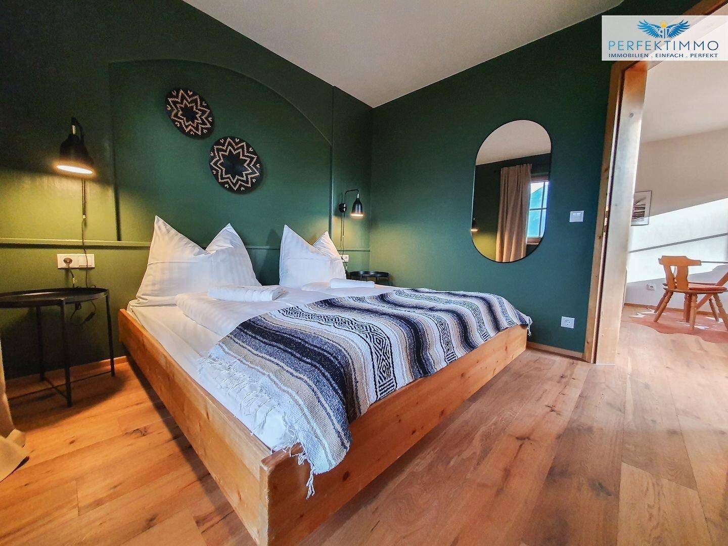 Schlafzimmer mit Holzbett, grüner Akzentwand und ovalem Spiegel.