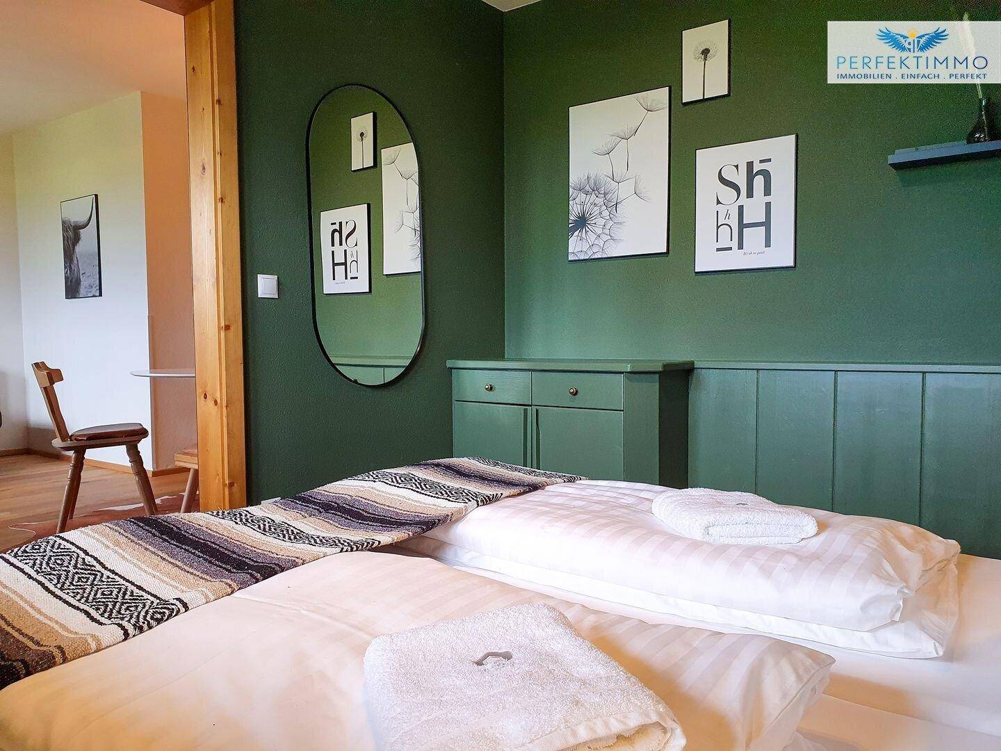 Schlafzimmer mit grüner Wand, modernen Bildern und einem ovalen Spiegel.