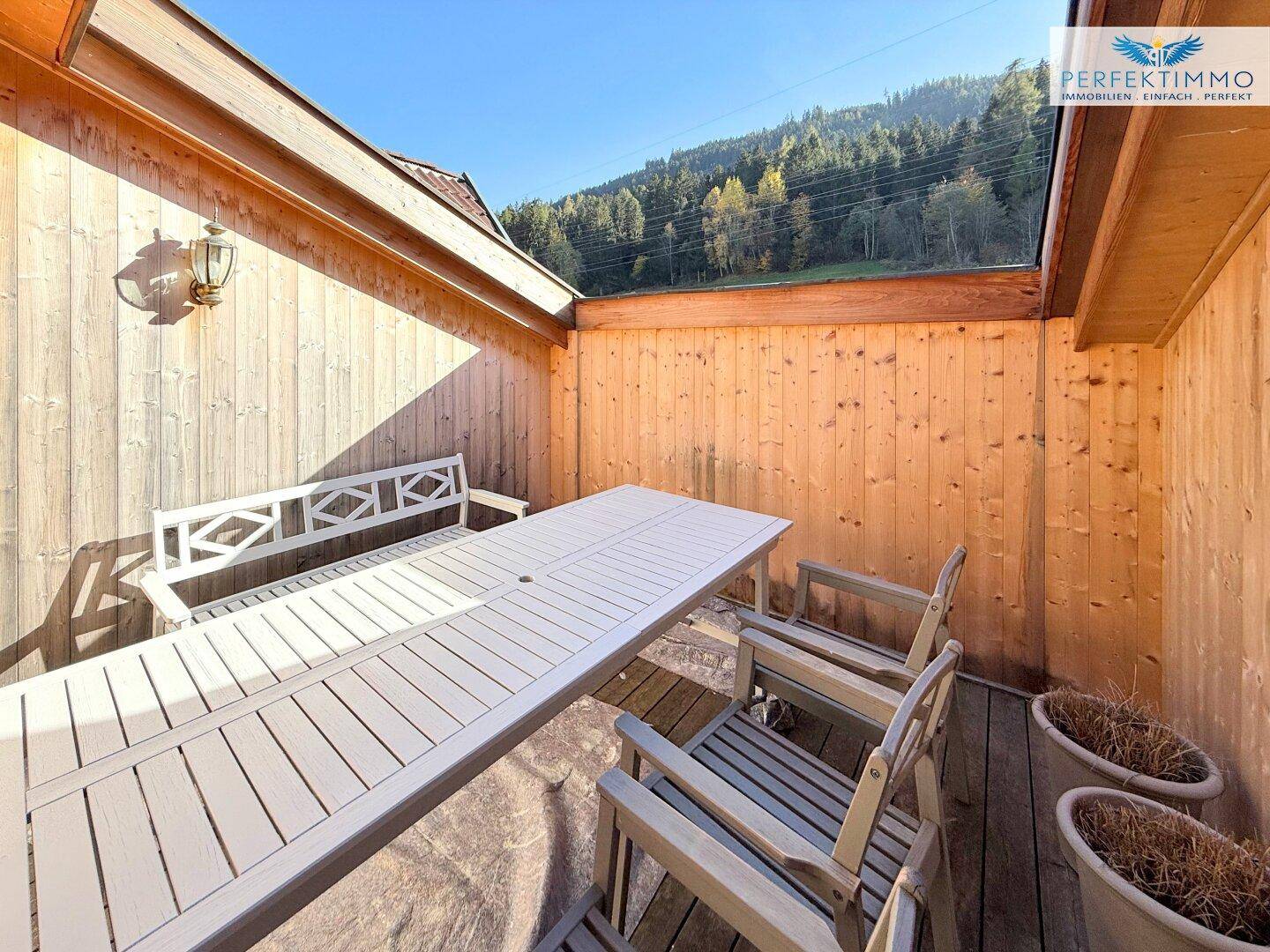 Sonniger Außenbereich mit Holzwänden, Esstisch und Stühlen, umgeben von grüner Natur.