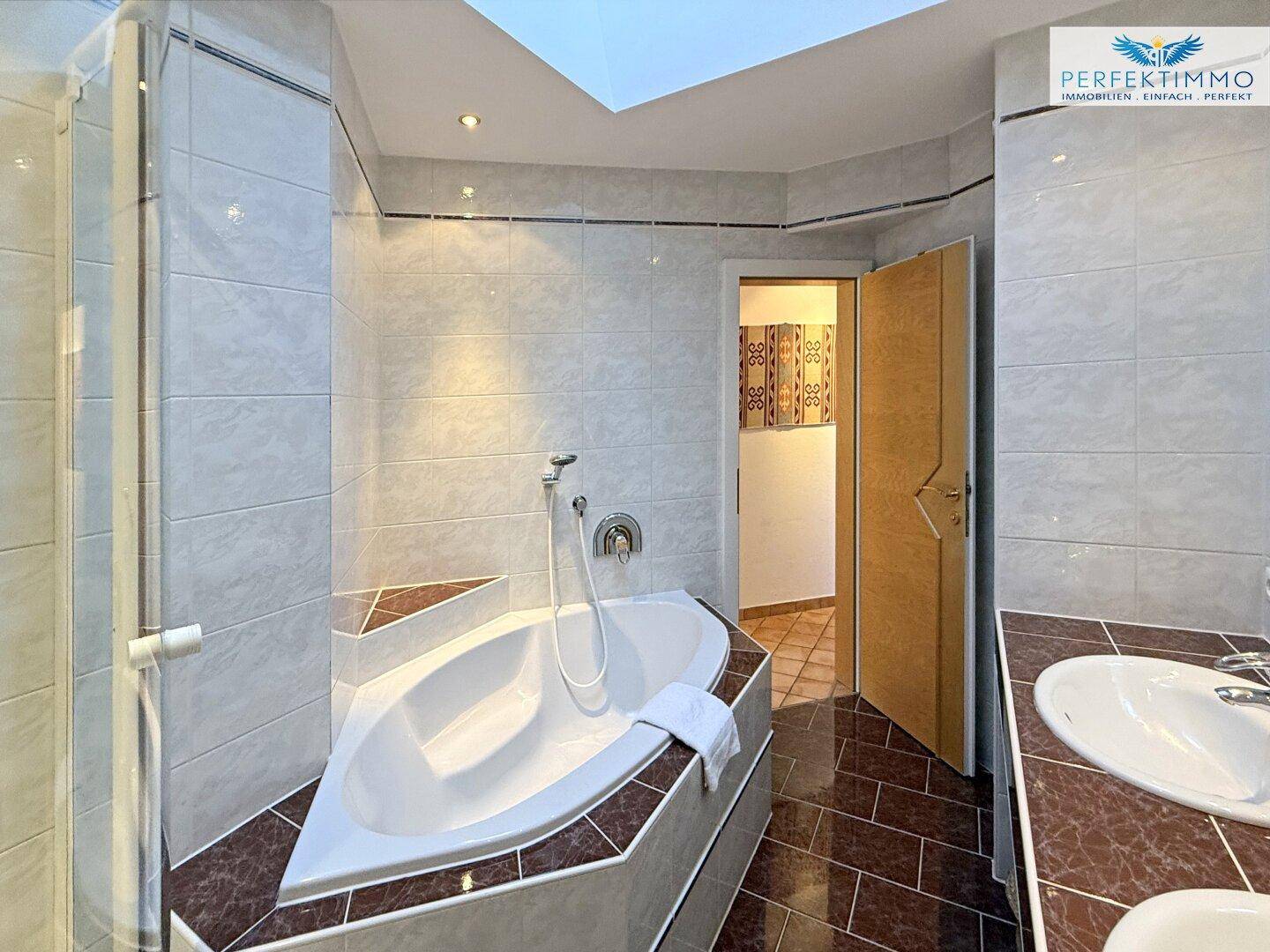 Badezimmer mit Eckbadewanne und Zugang zu einem weiteren Raum, hell gefliest.