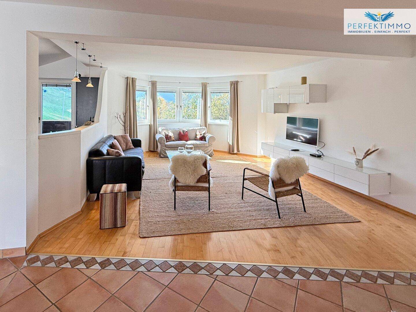 Heller Wohnbereich mit Sofas, TV-Möbeln und großen Fenstern, Übergang zu Holzboden.