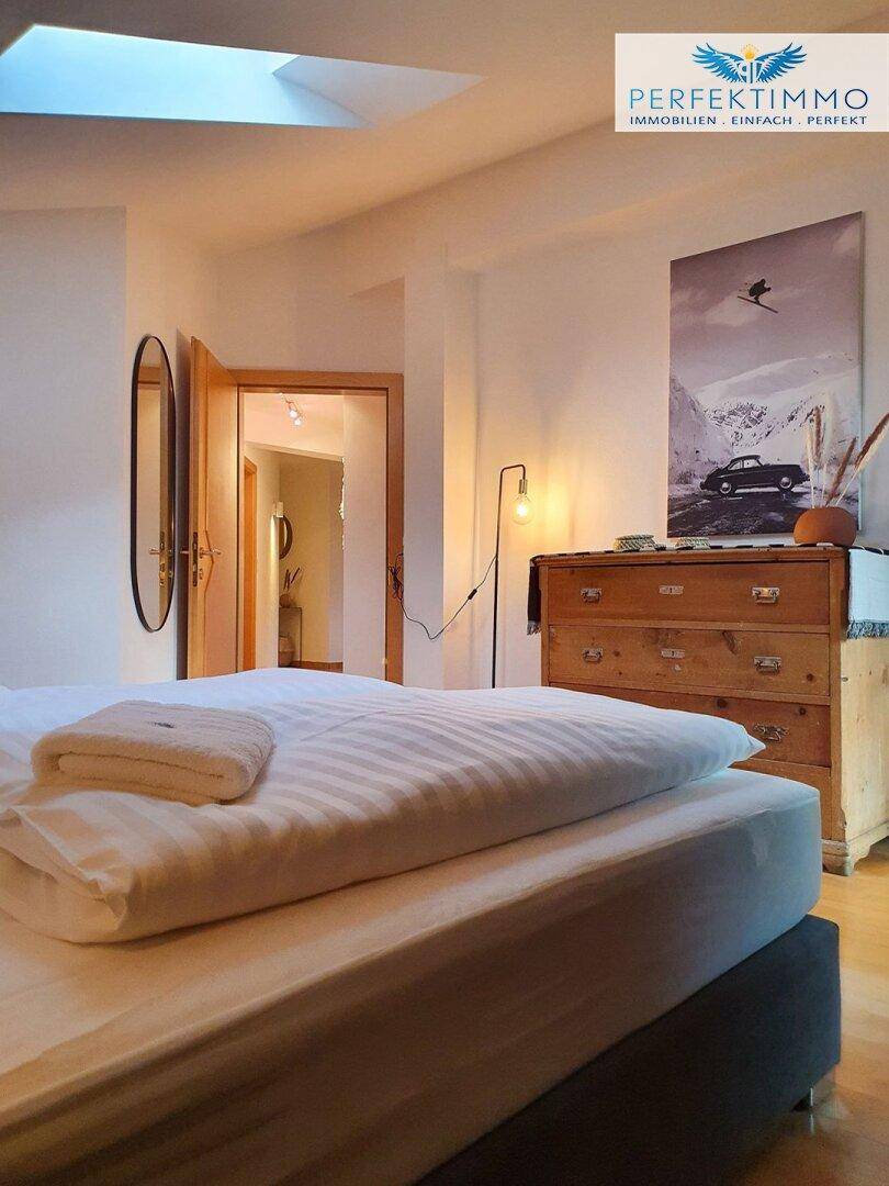 Gemütliches Schlafzimmer mit Bett, Kommode, Spiegel und einem Oberlicht für Tageslicht.