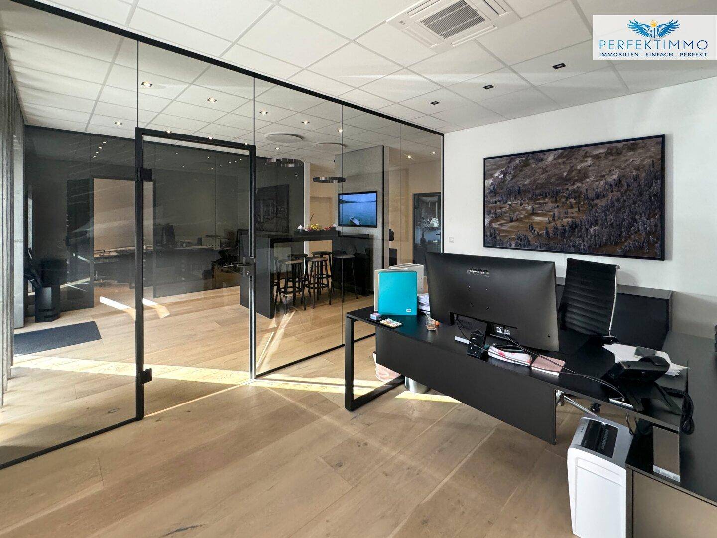 Geräumiges Büro mit Schreibtisch, Computer und transparenten Glaswänden zu anderen Bereichen.