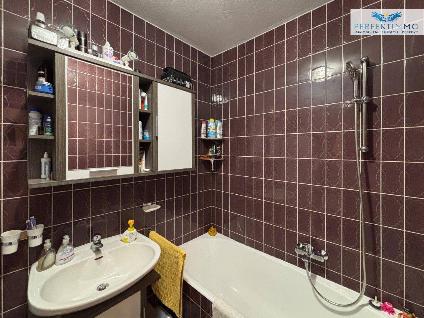 Badezimmer mit braunen Fliesen, Waschbecken und Badewanne mit Duschfunktion, funktional und gepflegt.