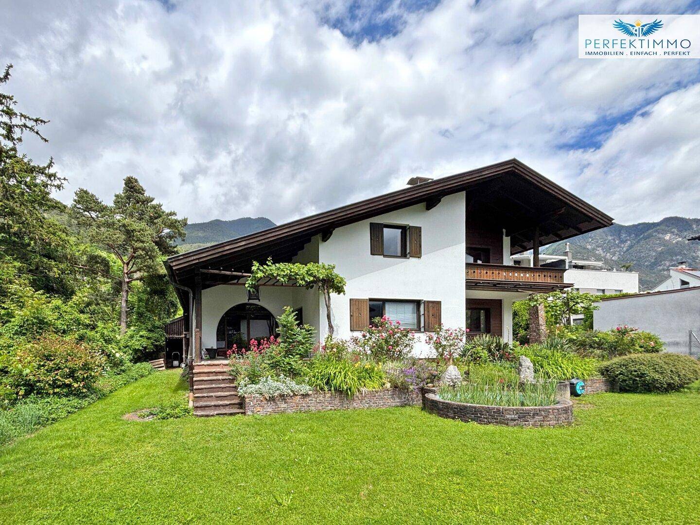 Traditionelles Einfamilienhaus mit großem Garten und Bergblick unter einem bewölkten Himmel.
