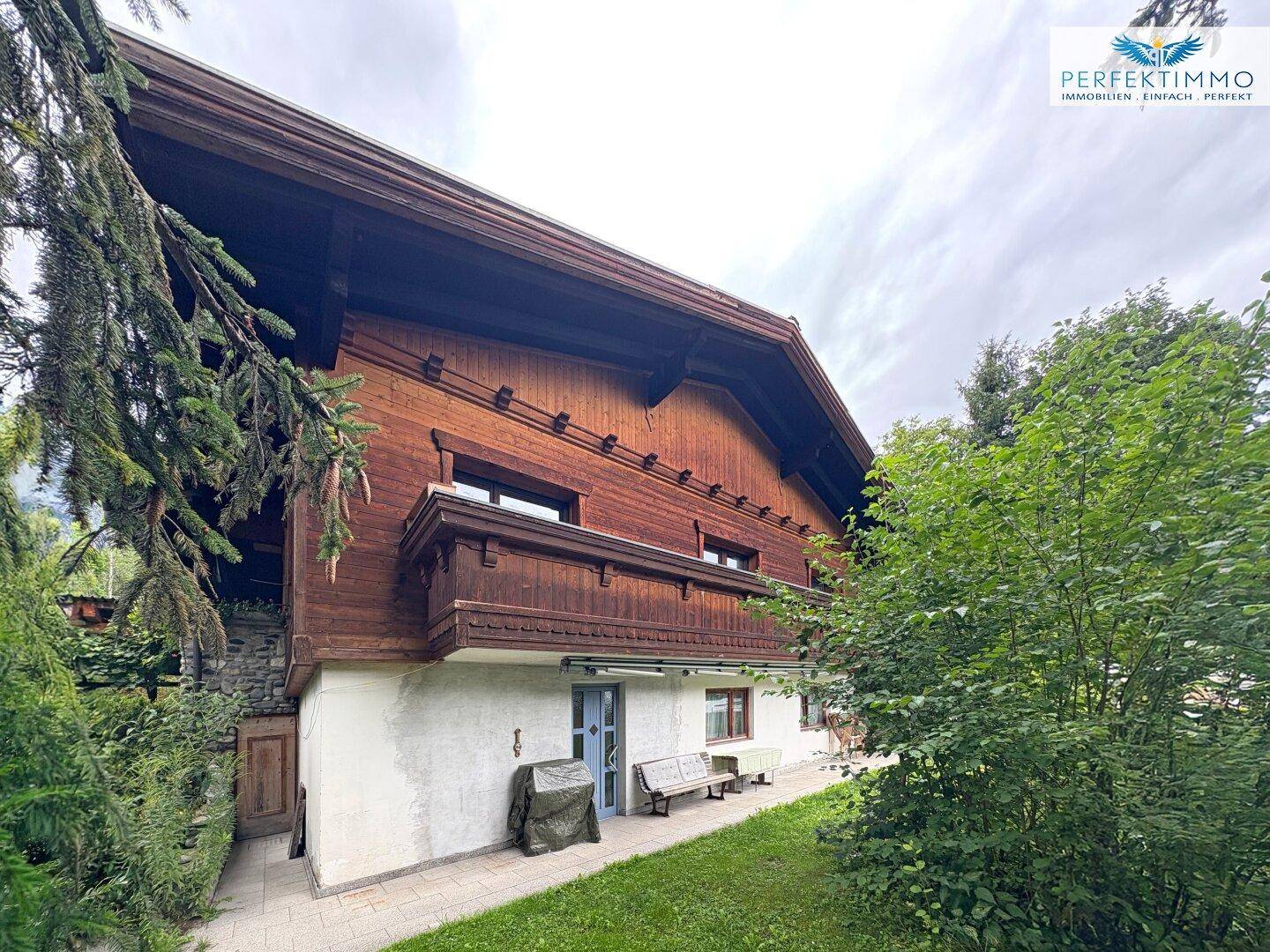 Traditionelles Holzhaus mit kleinem Balkon, gepflasterter Terrasse und umgeben von viel Grün.