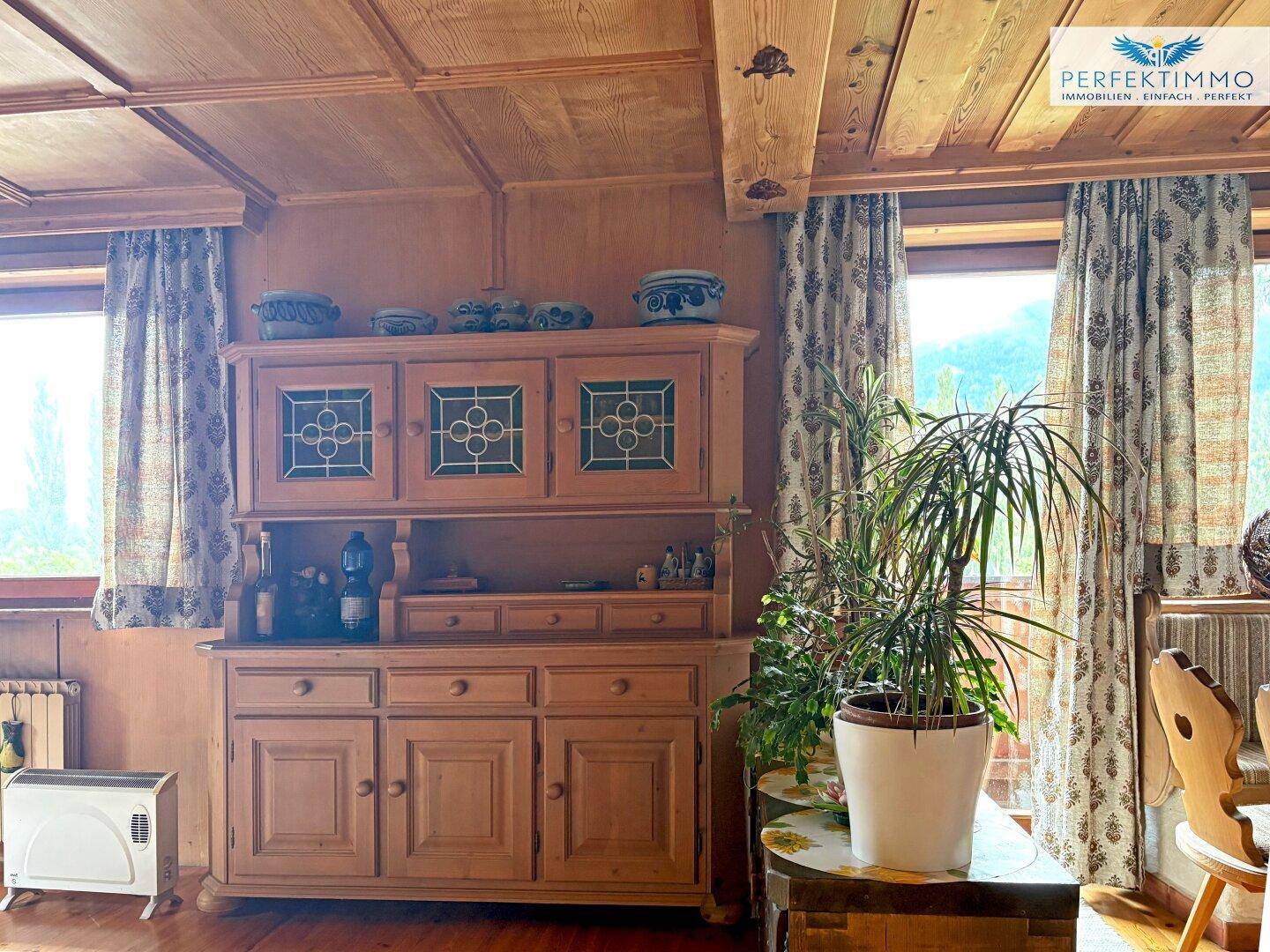 Rustikaler Wohnbereich mit traditionellem Holzschrank, Pflanzen und Blick ins Grüne.