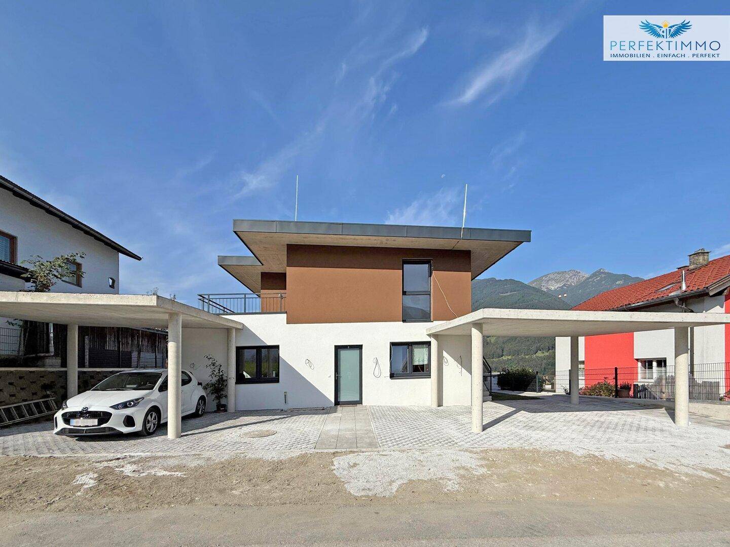 Ansicht des modernen Hauses mit zwei Carports und Bergpanorama im Hintergrund unter blauem Himmel.