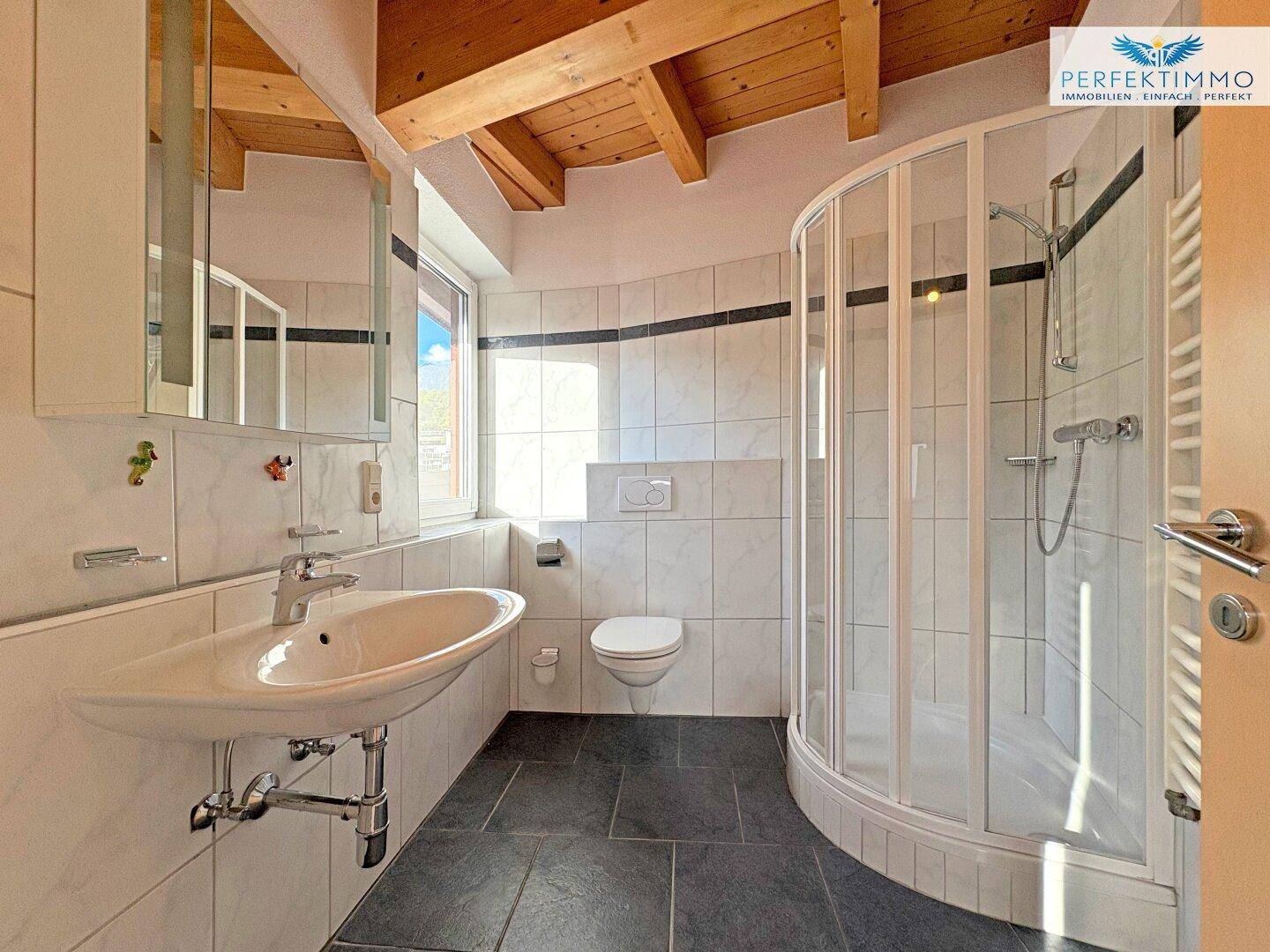 Helles Badezimmer mit Duschkabine, WC, Waschbecken und sichtbaren Holzbalken an der Decke.