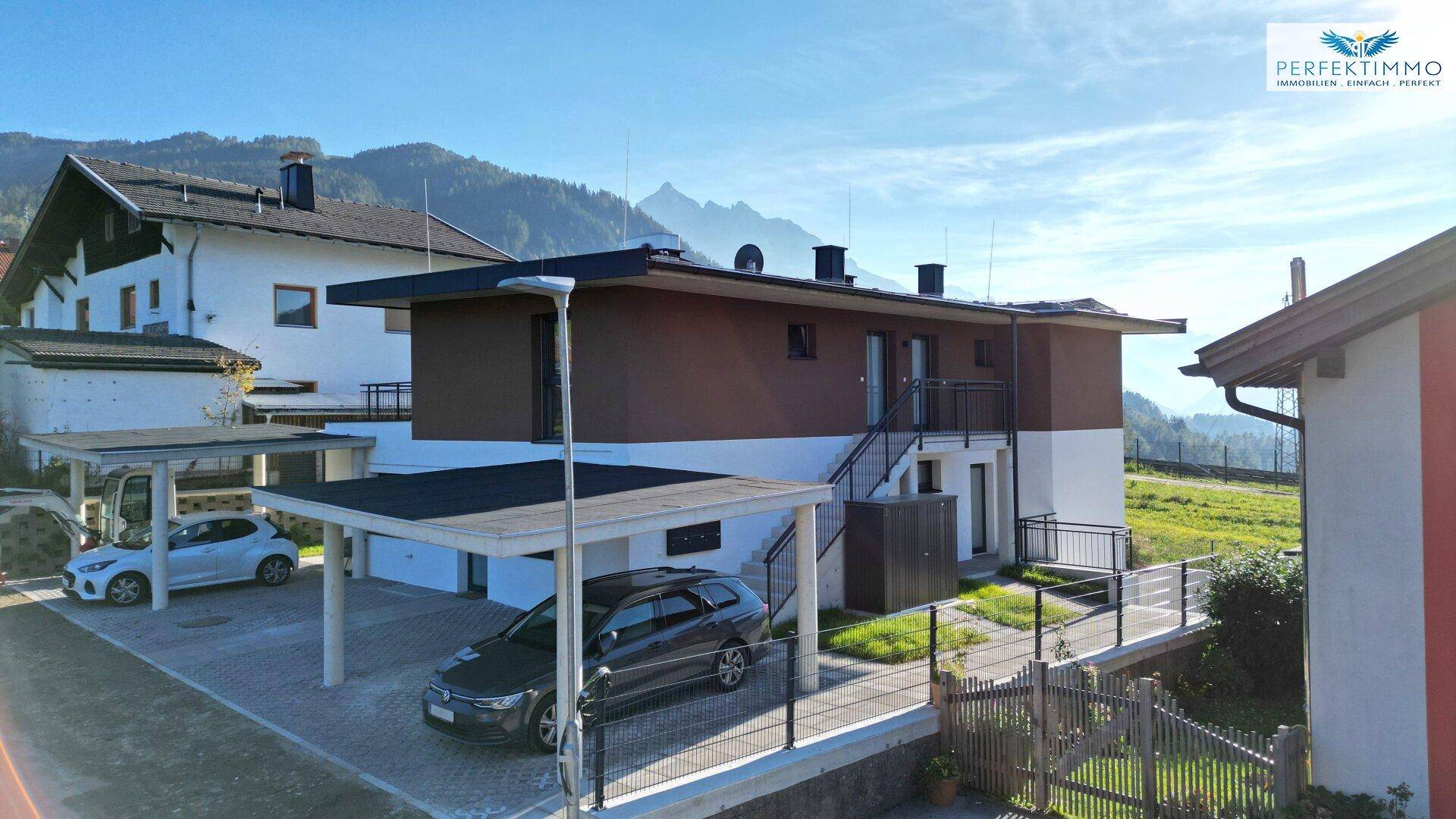 Modernes Wohngebäude mit Carports und Bergblick, umgeben von grüner Landschaft.