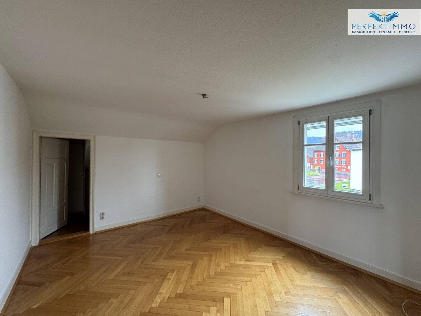 Heller Raum mit Fischgrätparkettboden und Fenster mit Blick auf die Umgebung.