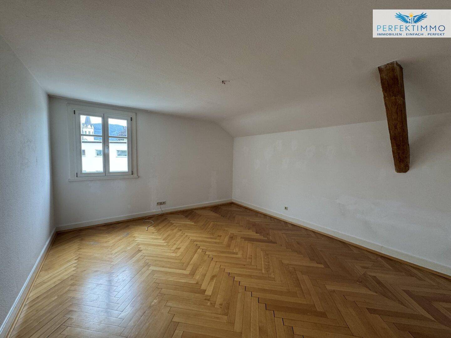 Geräumiger Raum mit Fischgrätparkettboden, Fenster und sichtbarem Holzbalken.