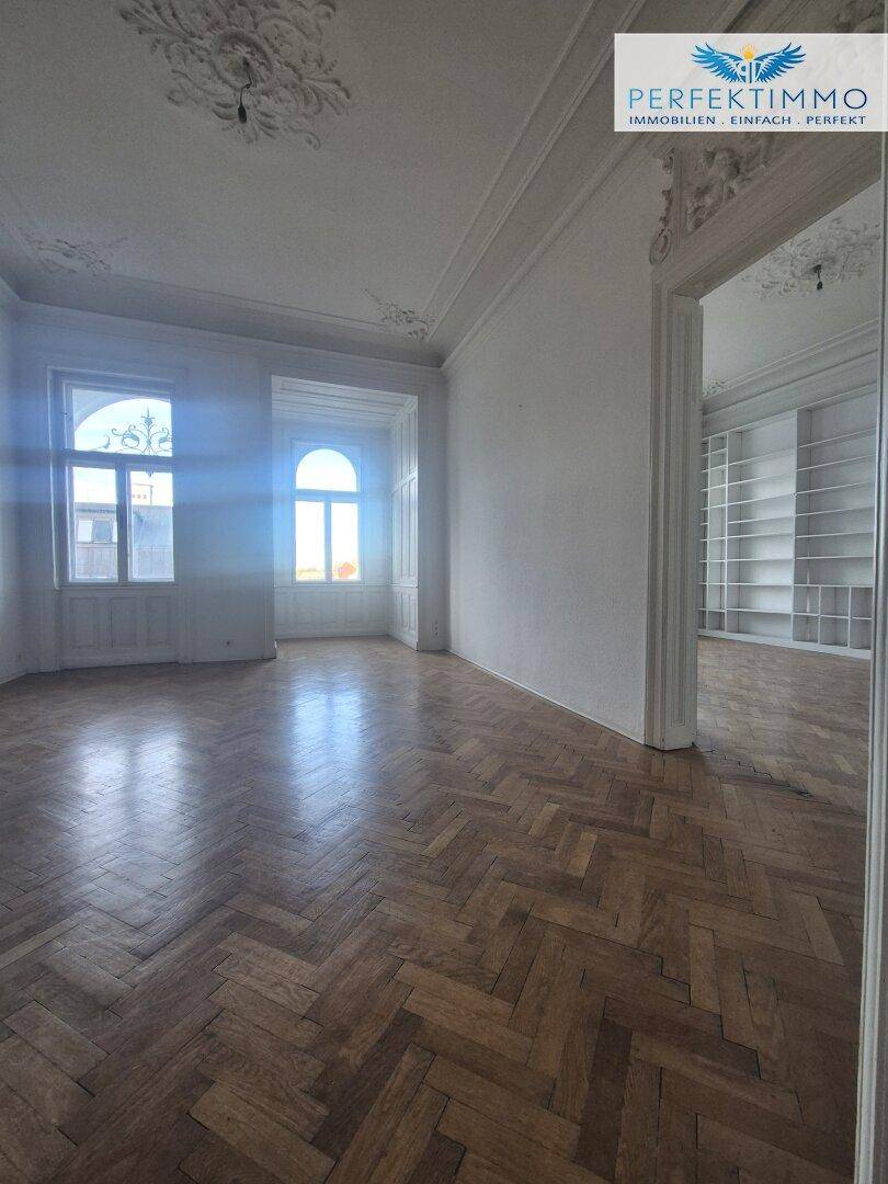 Großzügiges Wohnzimmer mit glänzendem Fischgrätparkett, hohen Decken und großen Fenstern für viel Tageslicht.